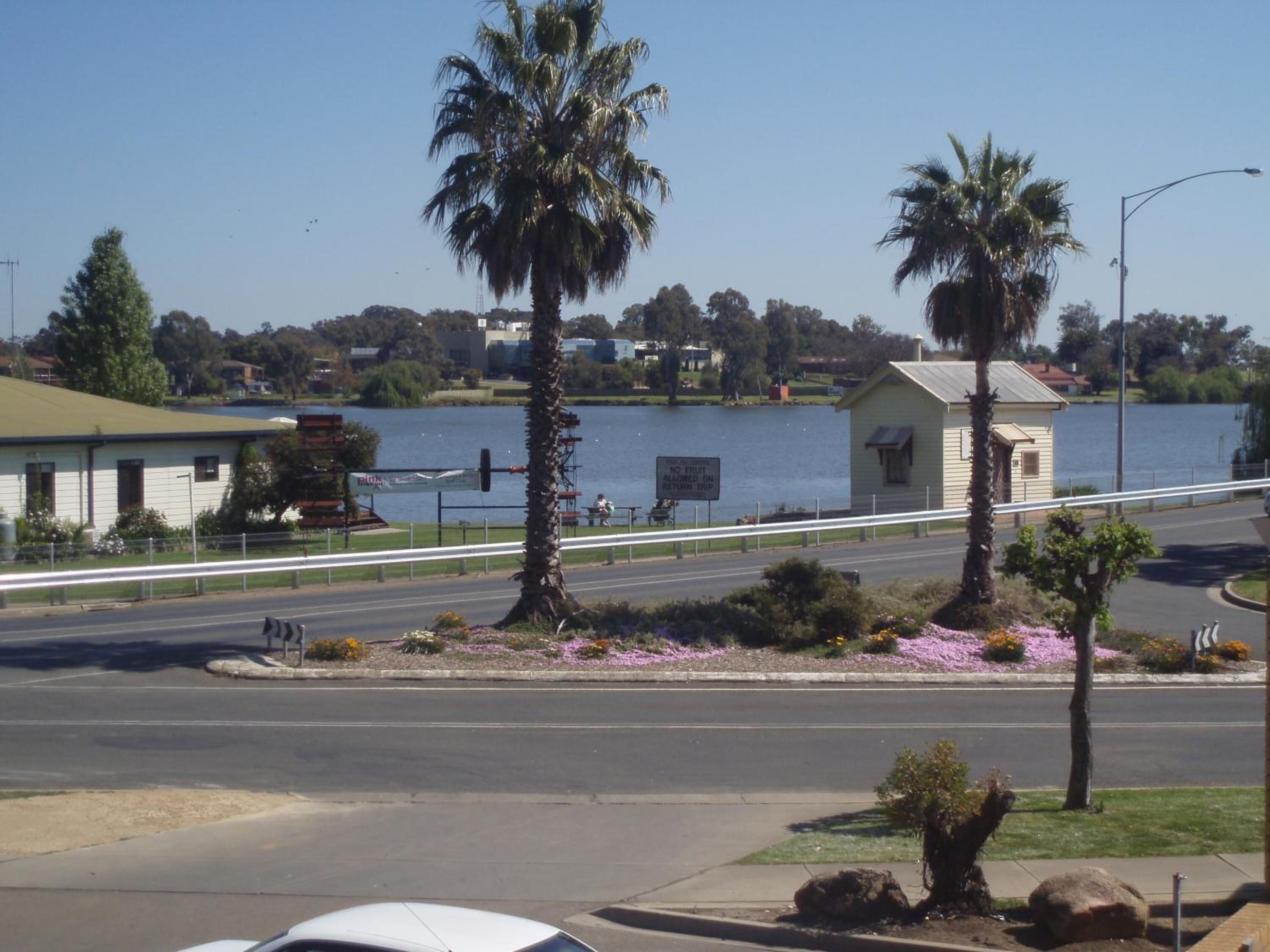 A road with a pedestrian bridge over a body of water, palm trees, and a building with a sign.