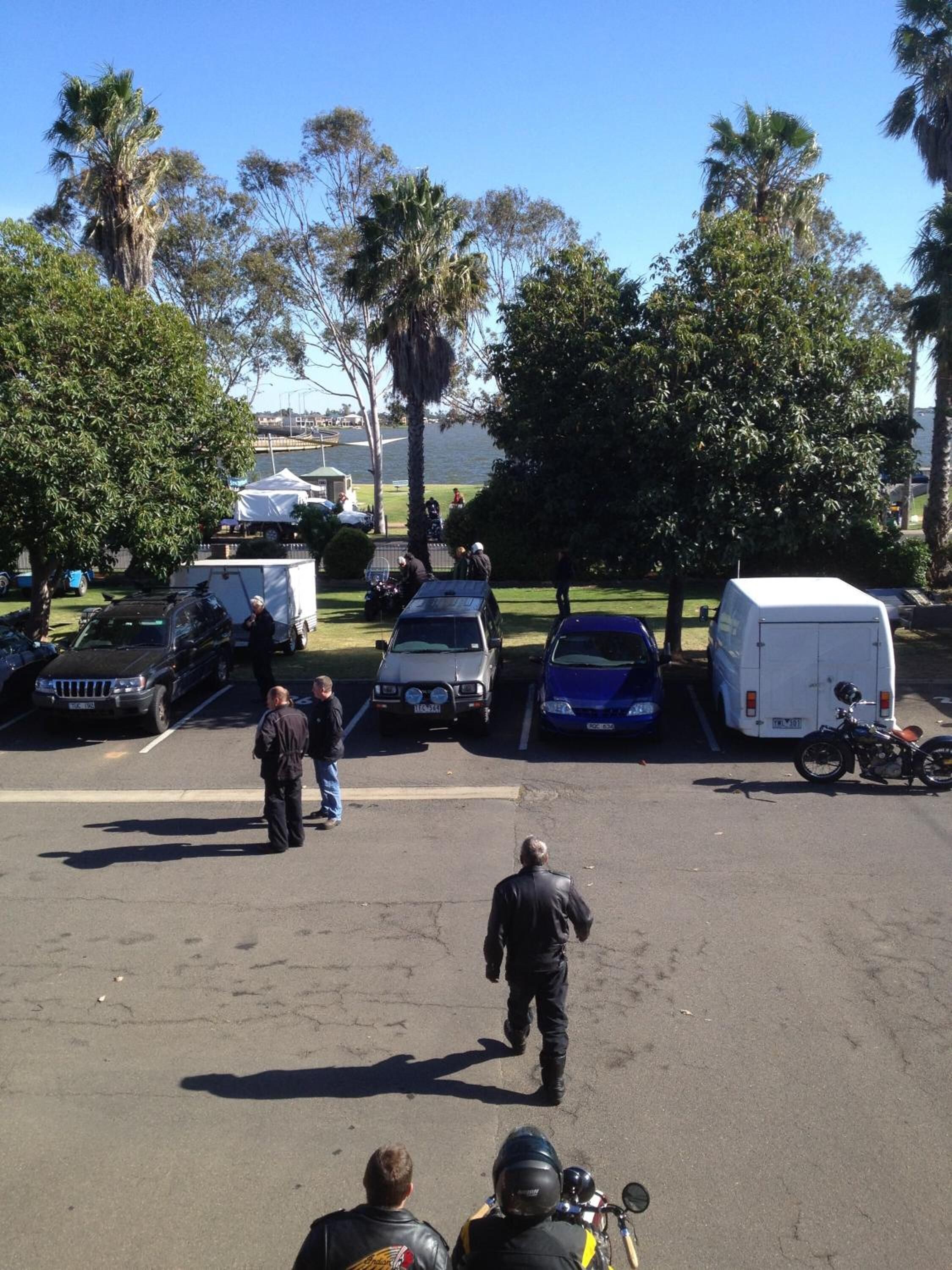 Un pàrquing amb diversos vehicles, incloent cotxes i motocicletes, i algunes persones passejant.