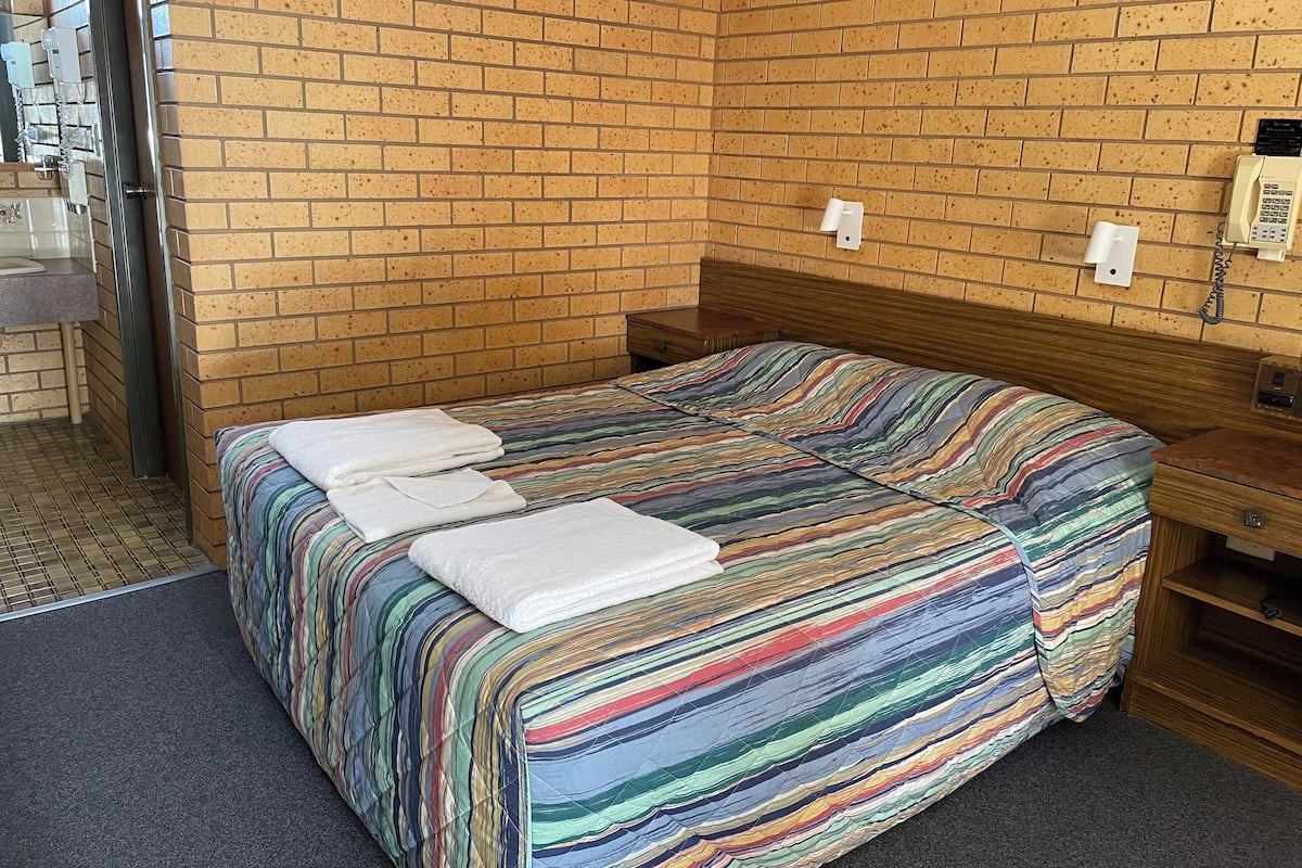 Una cama individual con una colcha a rayas de colores y dos toallas blancas. La habitación tiene una pared de ladrillo y un cabecero de madera. Hay una mesita de noche de madera con una lámpara y un teléfono en la pared.