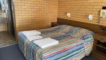 Una cama individual con una colcha a rayas de colores y dos toallas blancas. La habitación tiene una pared de ladrillo y un cabecero de madera. Hay una mesita de noche de madera con una lámpara y un teléfono en la pared.
