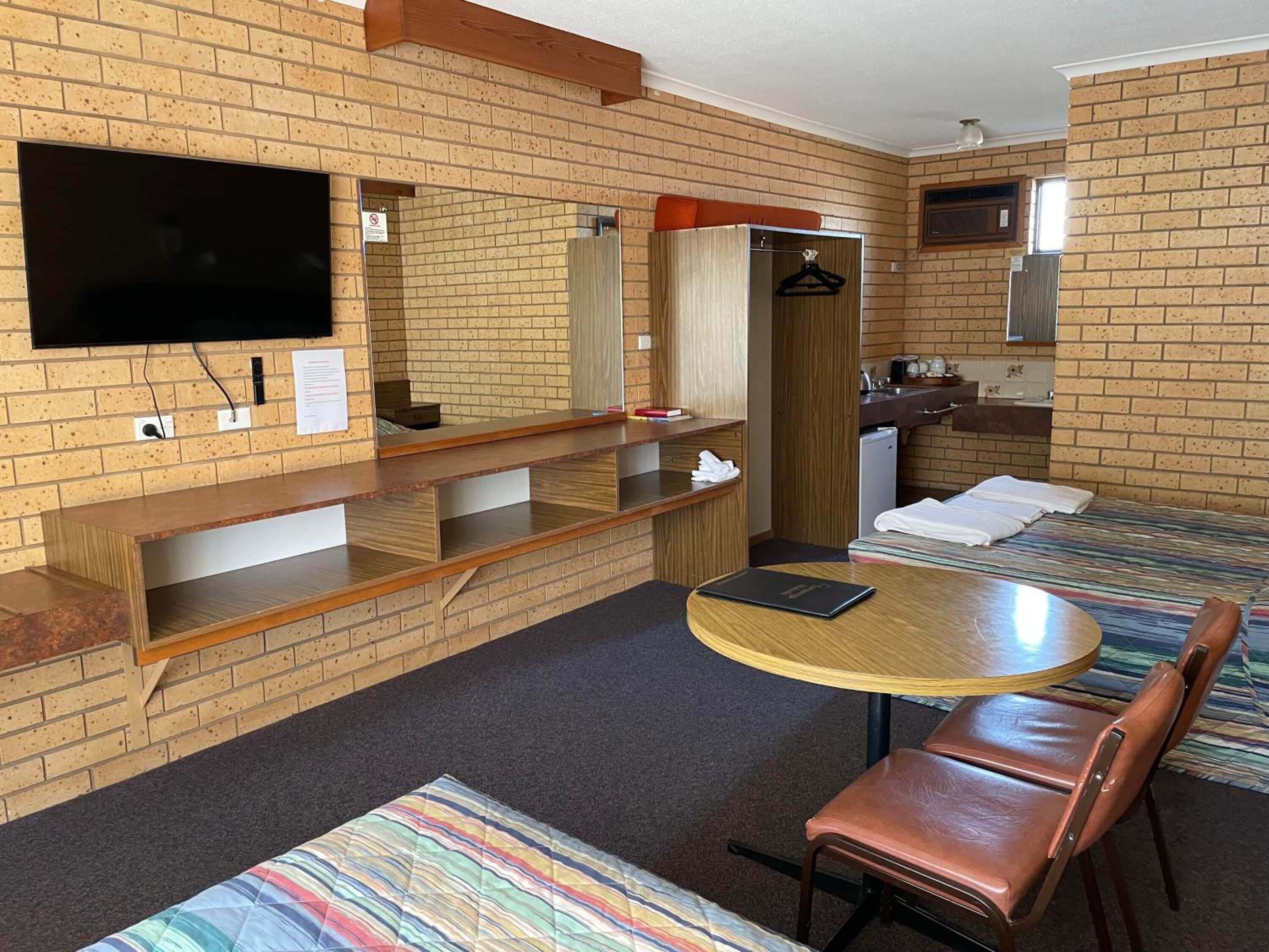 A hotel room with a brick wall, a flat-screen TV mounted above a wooden desk, a round table with a chair, a bed with a colorful striped bedspread, and a small kitchenette area.