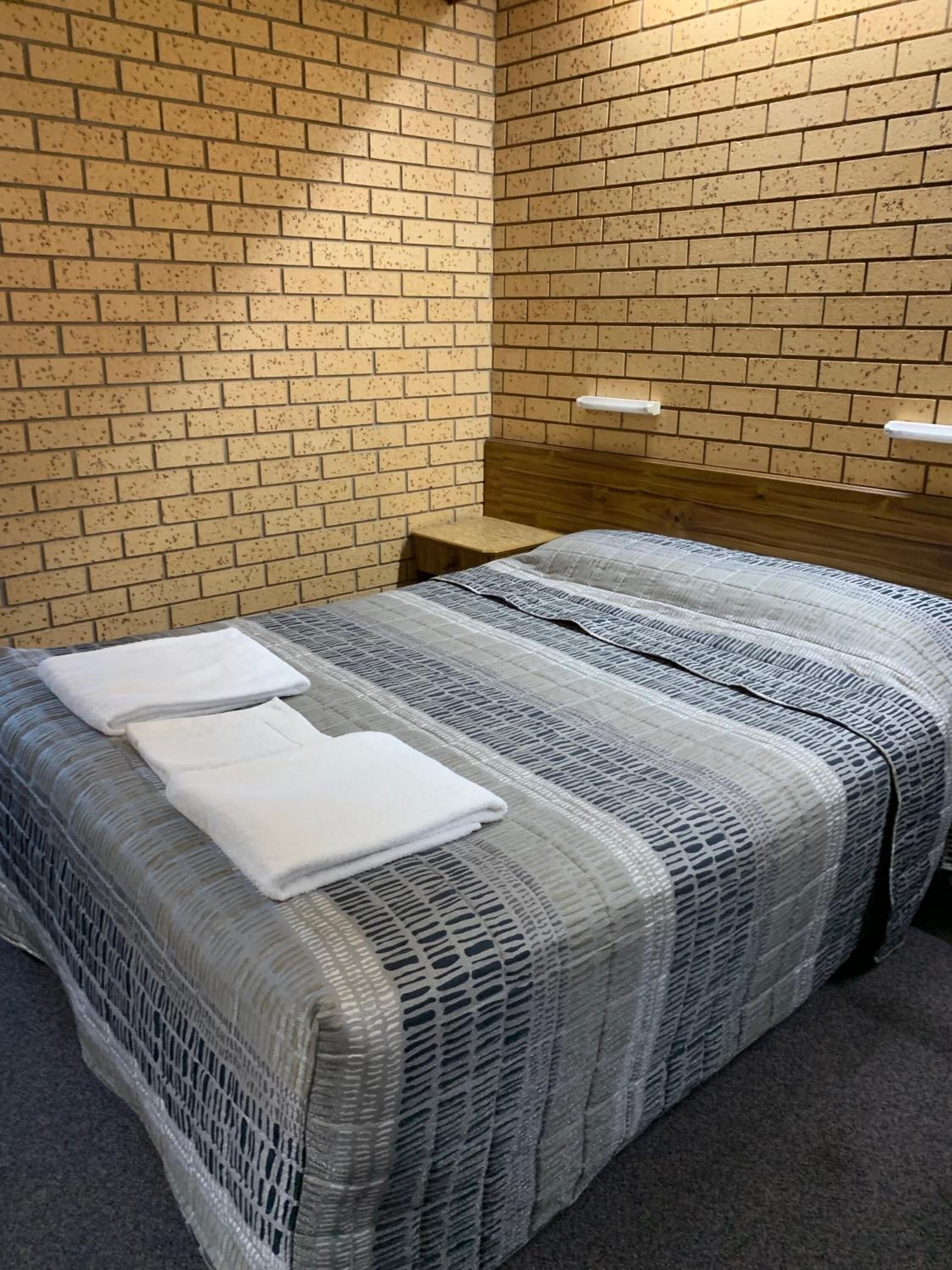A single bed with a grey and white striped bedspread, two white towels, and a wooden headboard in a room with brick-patterned walls.