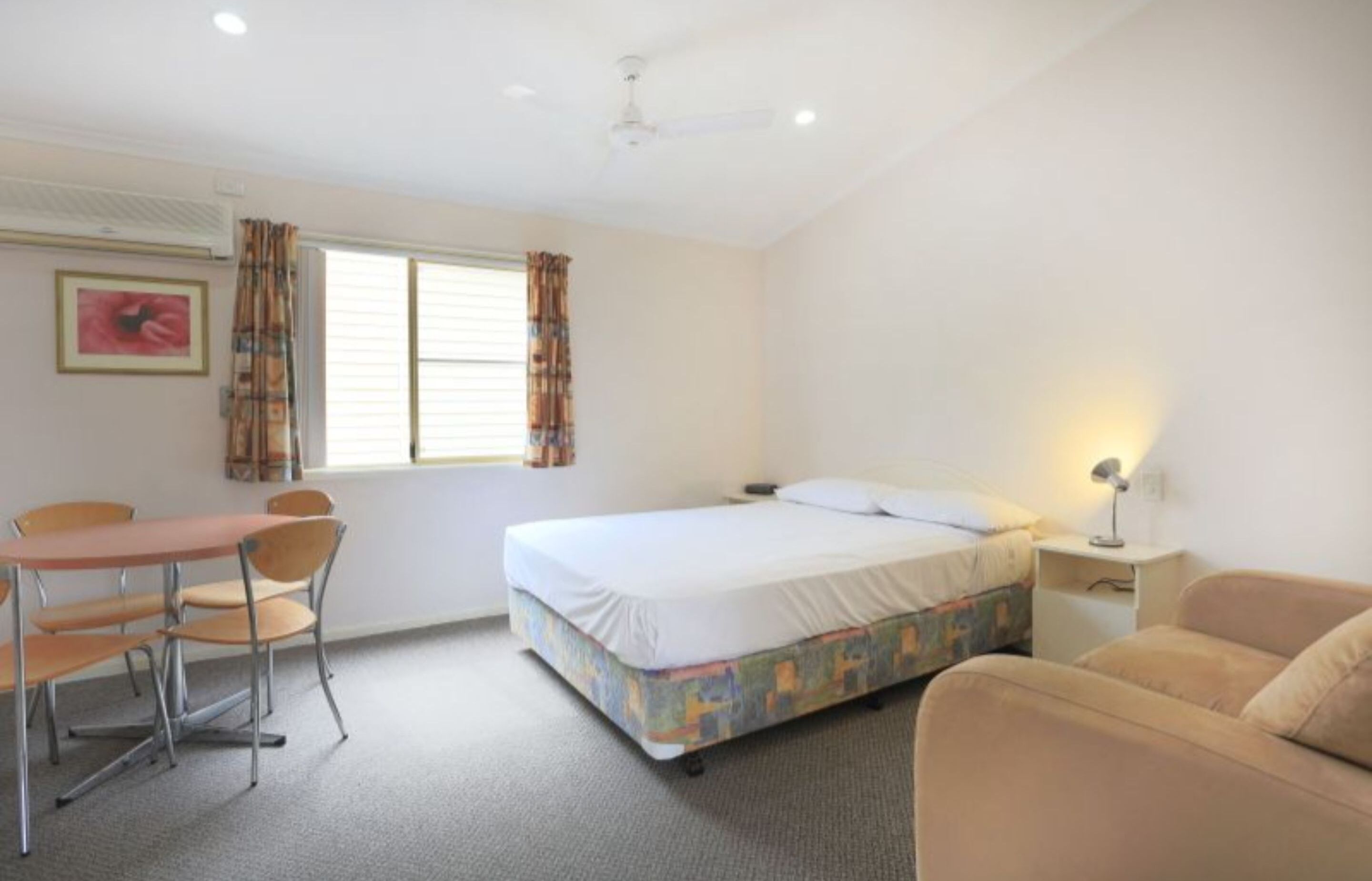 A bedroom with a bed, a desk with chairs, a bedside table with a lamp, and a window with curtains.