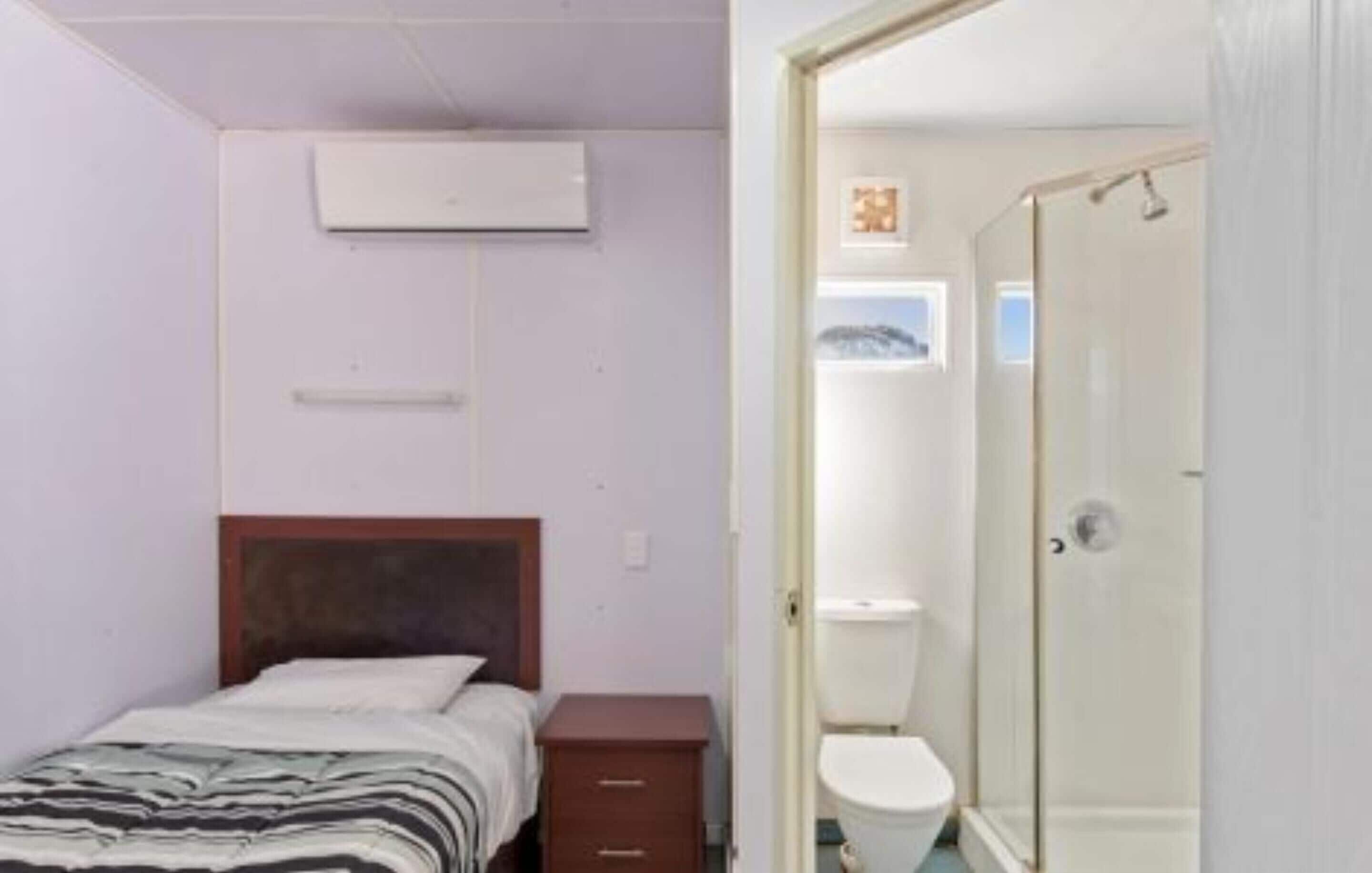 A small, clean room with a bed, a wooden nightstand, a small cabinet, and a shower area.