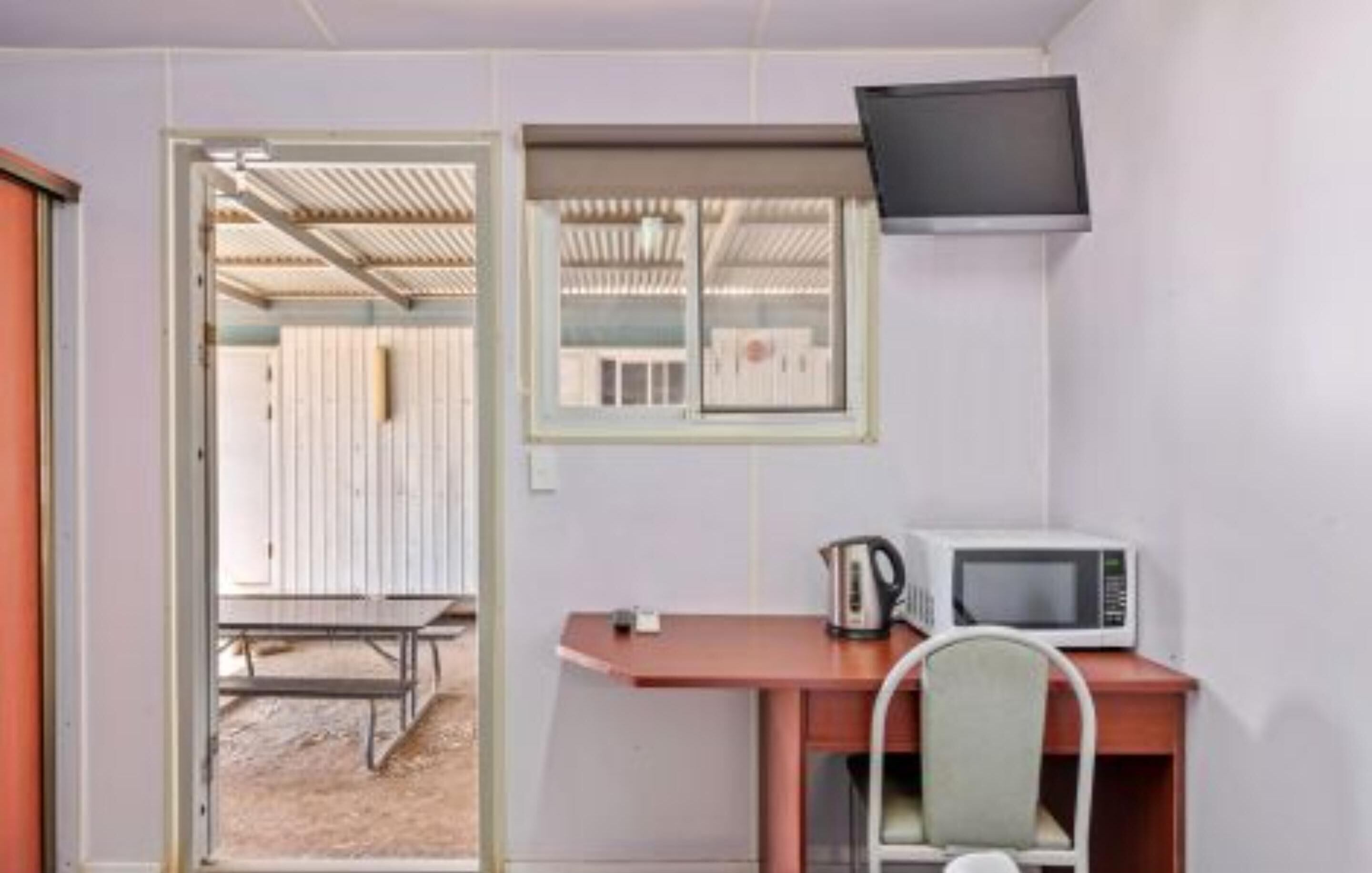 A small room with a desk, a chair, a television mounted on the wall, and a window with blinds.