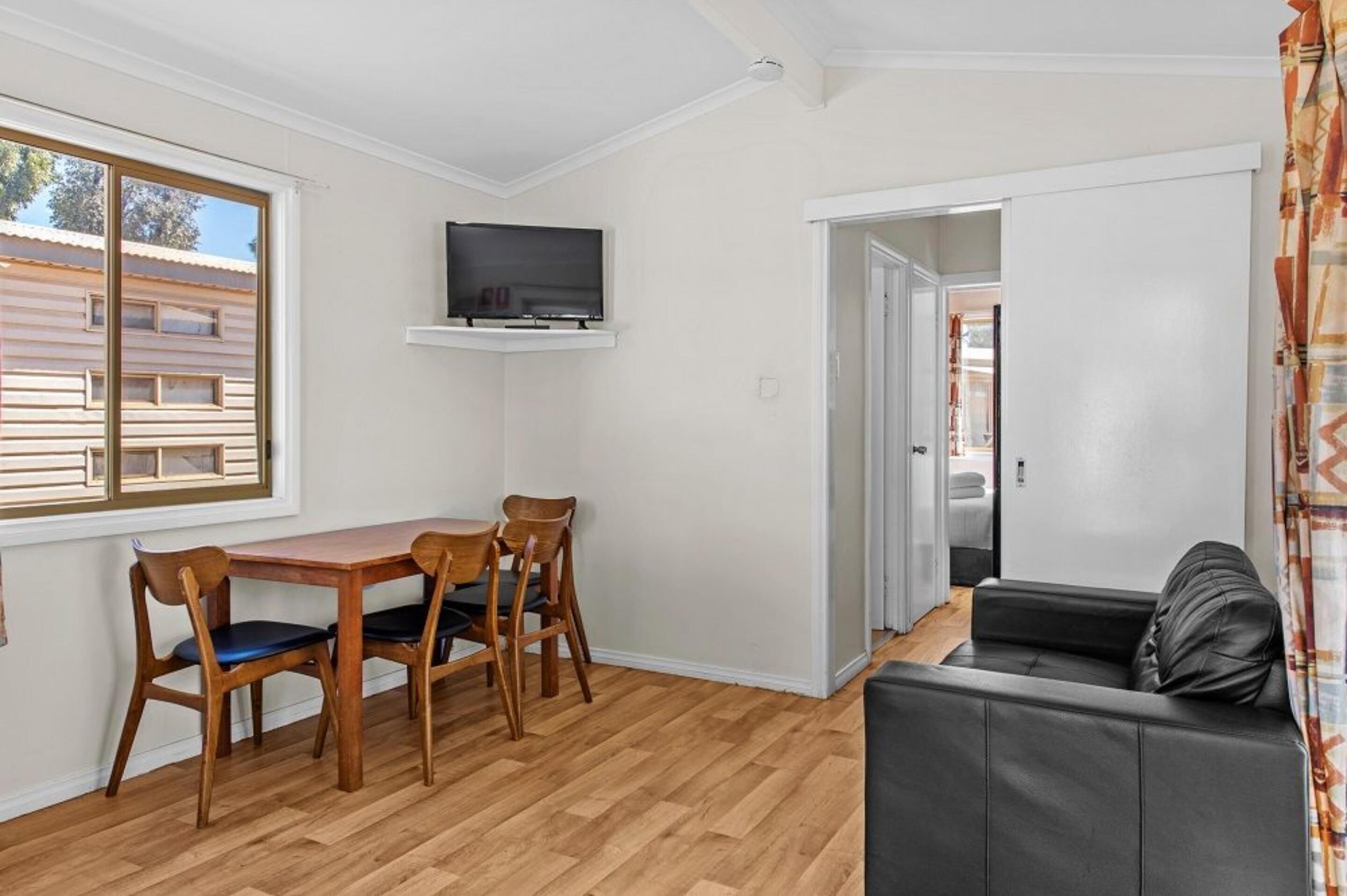 Un espace de vie compact comprenant une table à manger en bois, deux chaises, un canapé en cuir noir et un téléviseur fixé au mur.