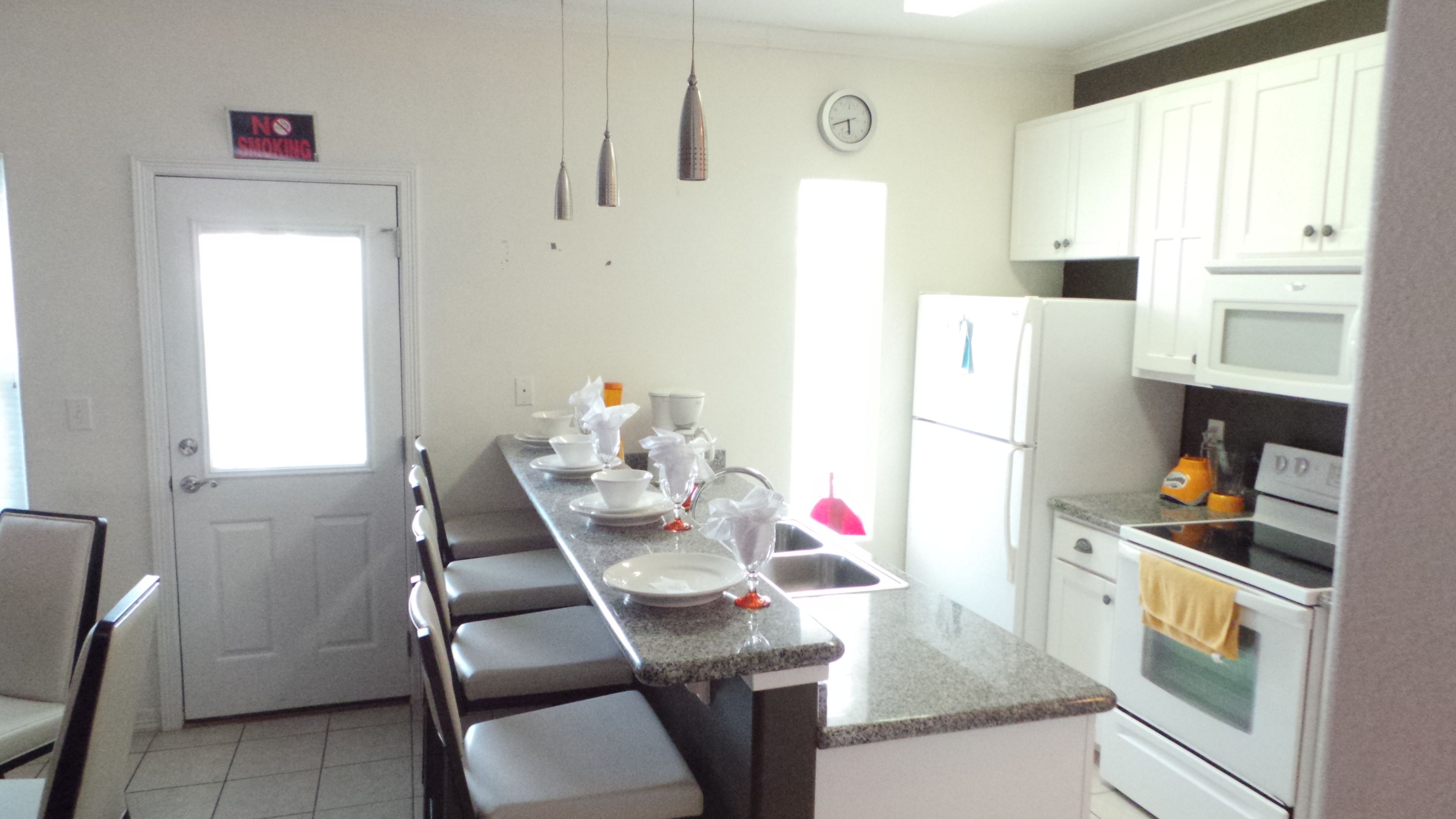 A kitchen with white cabinets, a refrigerator, a microwave, and a sink. There are chairs and a dining table with dishes and glasses.