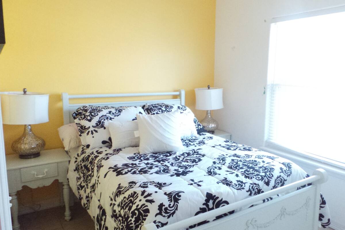 A bed with a white frame and a patterned bedspread, two bedside lamps, a nightstand with a drawer, and a window with blinds.