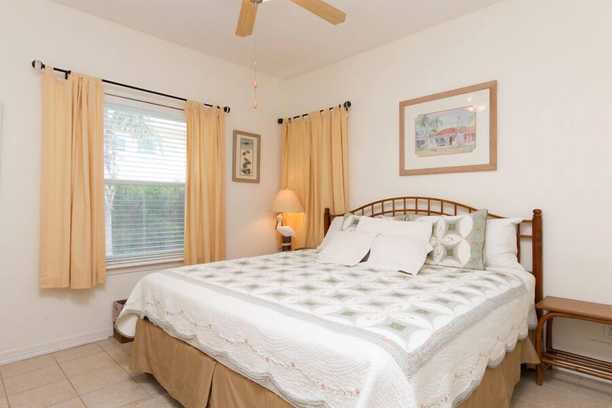 A bedroom with a bed, curtains, a ceiling fan, a bedside table, and a wall-mounted lamp.