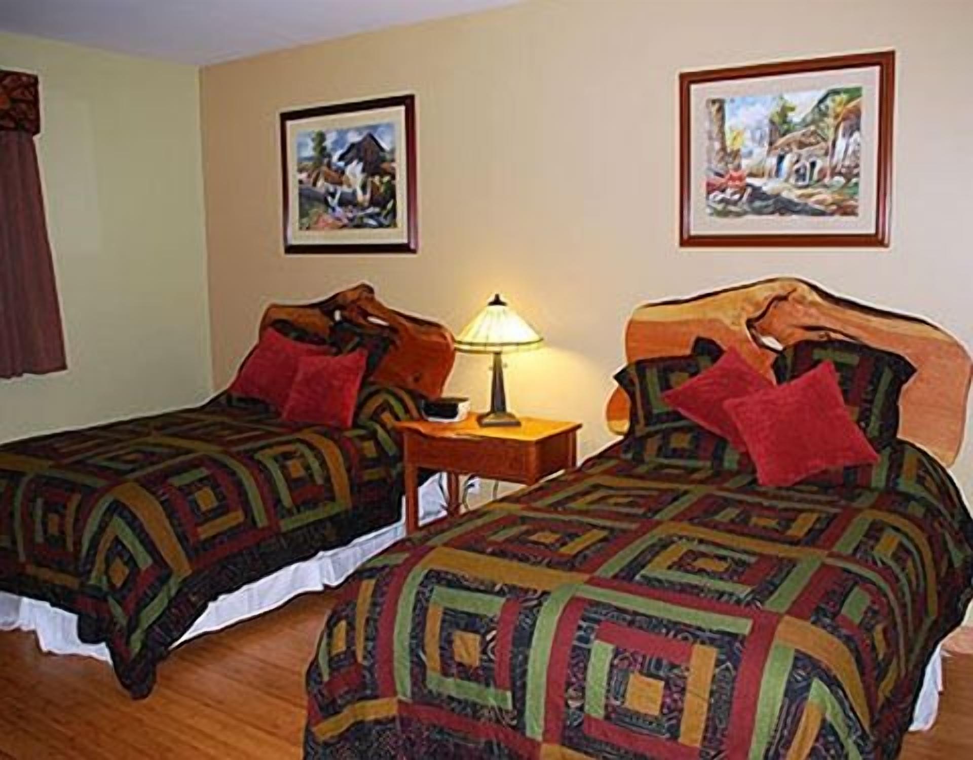 Une chambre avec deux lits, chacun recouvert d’une couvre-lit à motifs et agrémenté d’un oreiller rouge. Une table de chevet avec une lampe se trouve entre les deux lits. Deux tableaux encadrés sont accrochés au mur, au-dessus des lits.