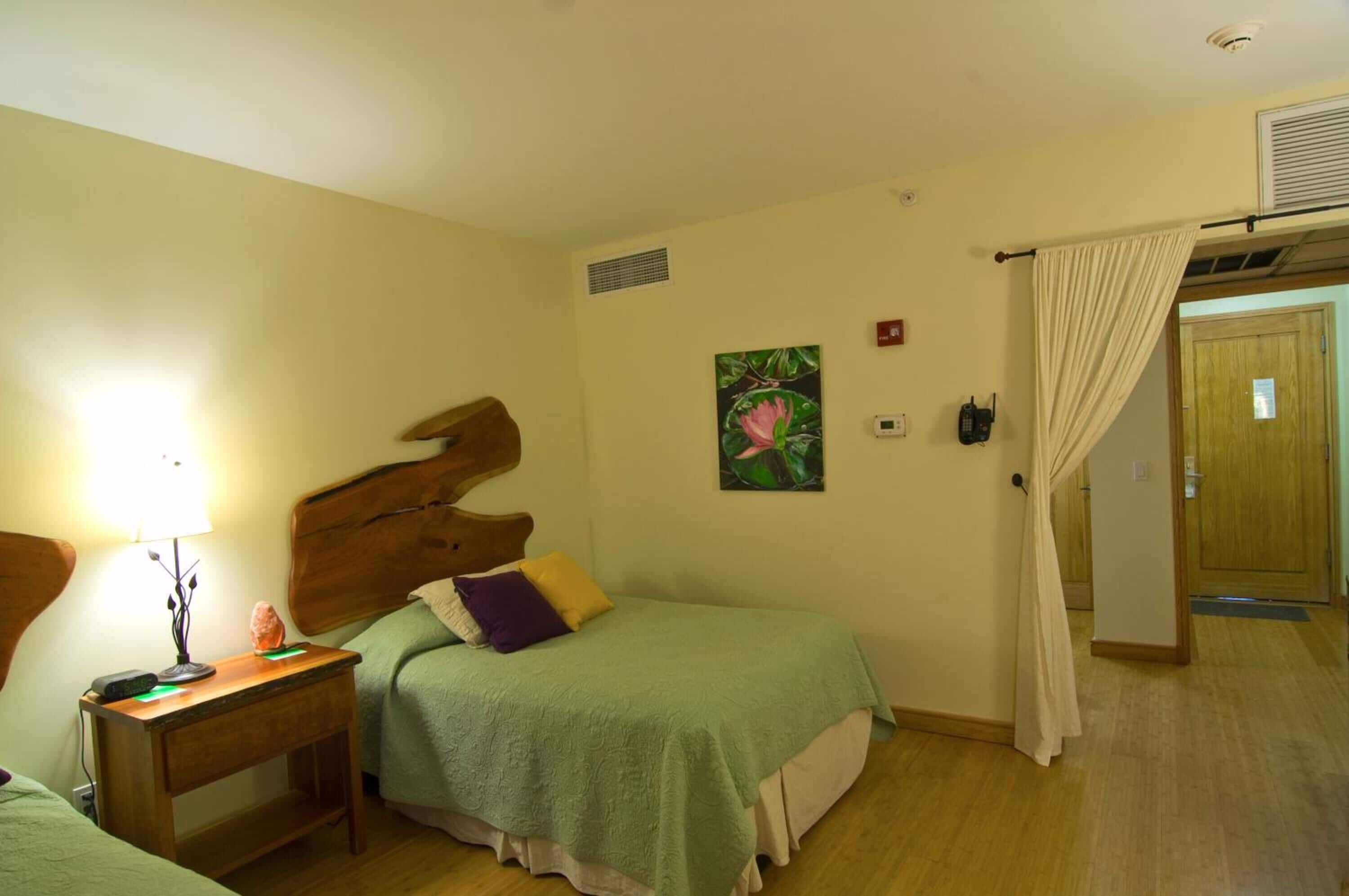 A bedroom with two beds, a wooden headboard, a bedside table with a lamp, a painting on the wall, and a door with a curtain.