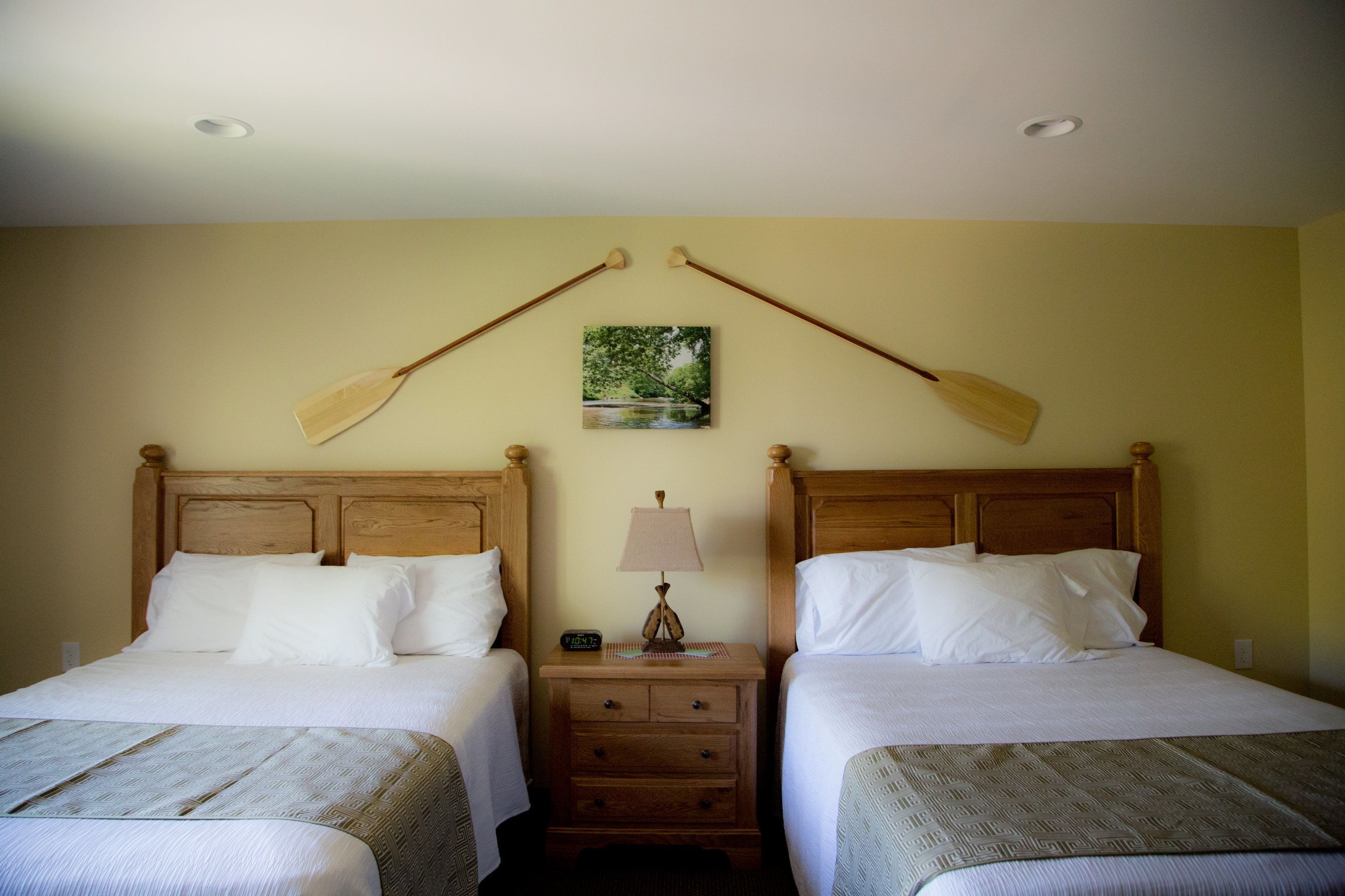 A hotel room with two single beds, wooden headboards, a nightstand with a lamp, and a painting of a landscape.