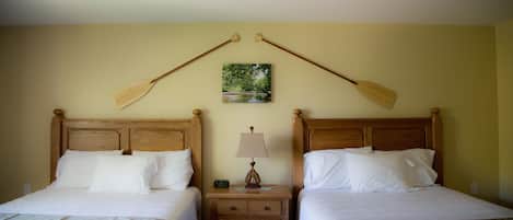 A hotel room with two single beds, wooden headboards, a nightstand with a lamp, and a painting of a landscape.