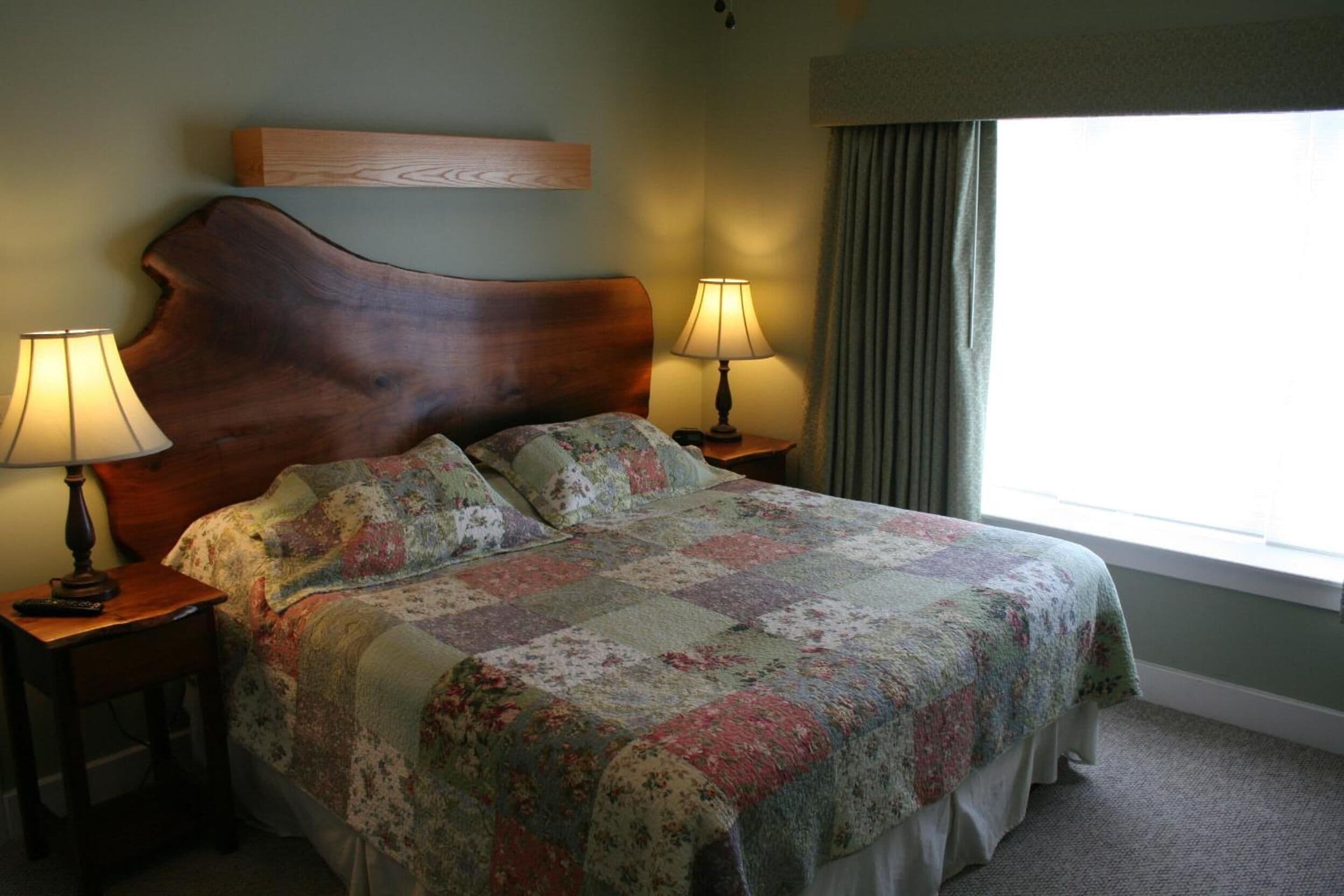A neatly made bed with a floral quilt, two bedside lamps, a wooden headboard, and a window with curtains.