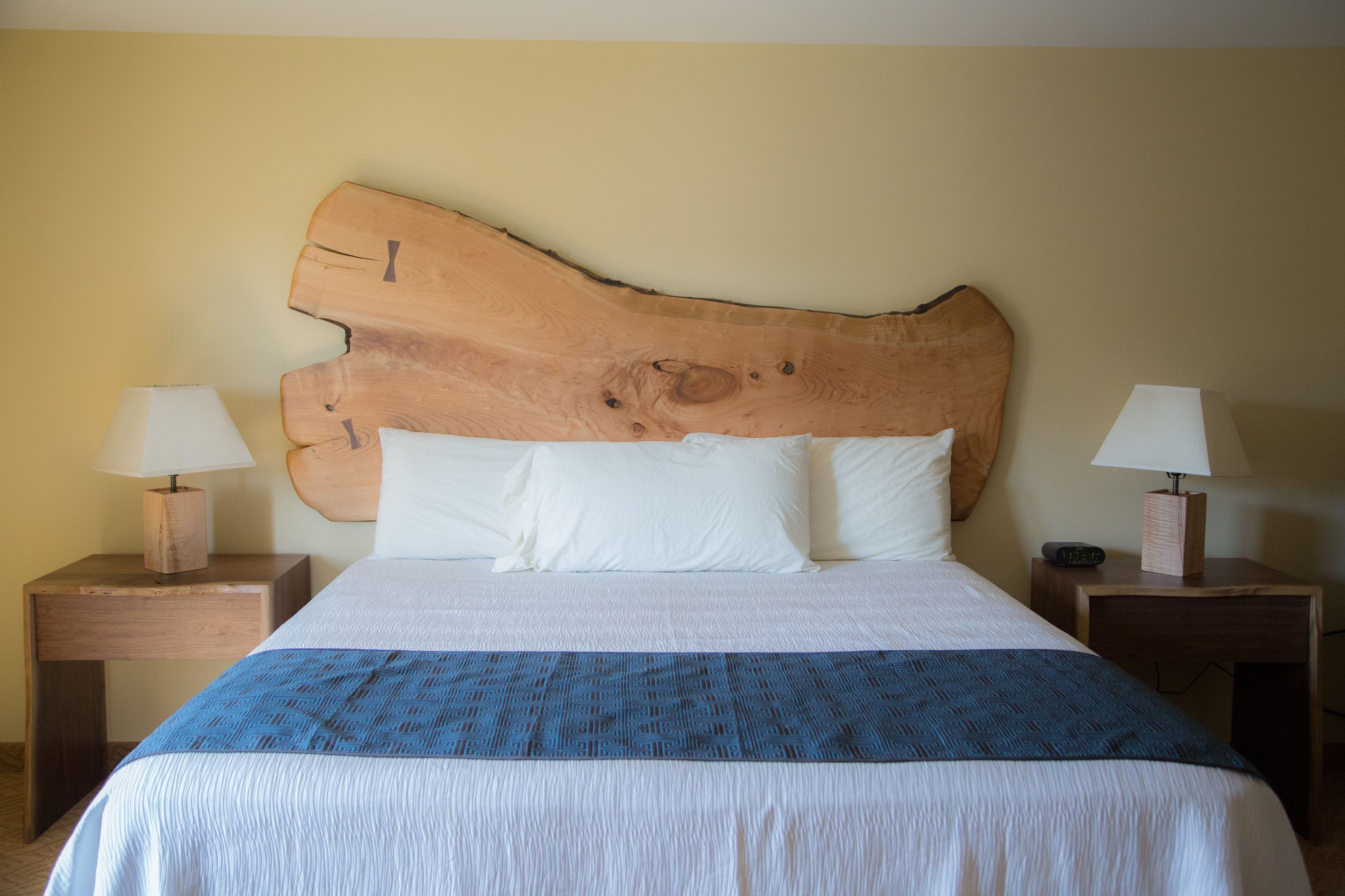 Un lit avec une tête de lit en bois, une literie blanche et une couverture bleue. Il y a deux tables de chevet, chacune avec une lampe, et une horloge sur l’une d’elles.