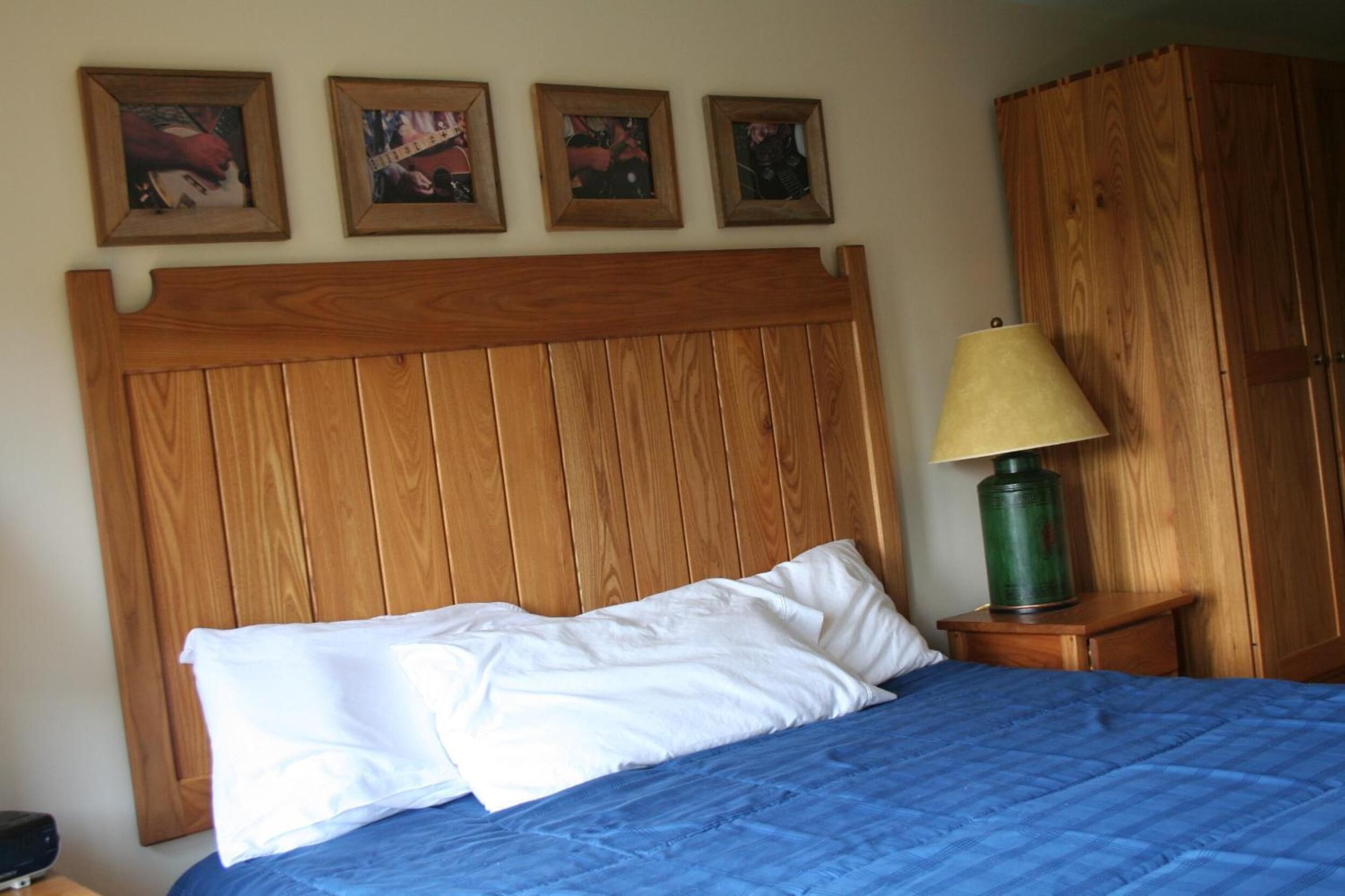 Ein Schlafzimmer mit einem hölzernen Kopfteil, einer blauen Bettdecke, weißen Kissen, einer grünen Lampe auf einem Nachttisch aus Holz und vier gerahmten Bildern an der Wand.