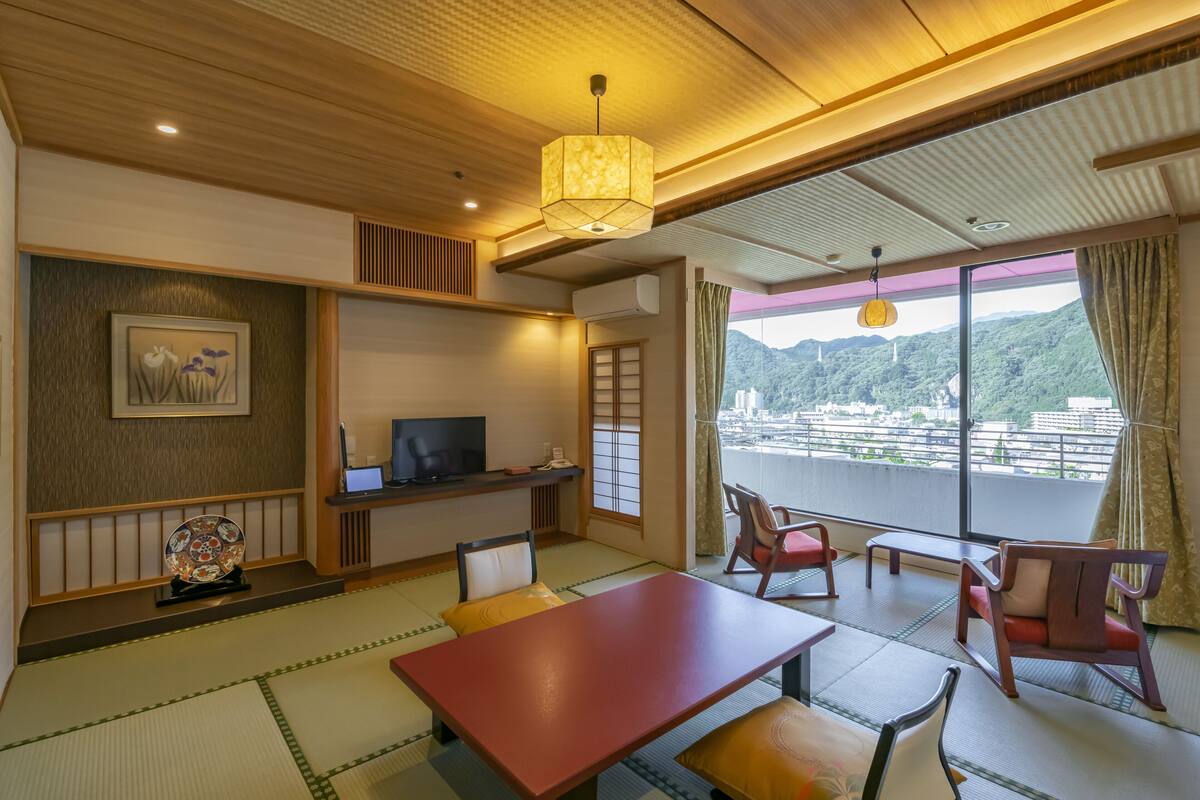 A traditional Japanese-style room with tatami flooring, a low table, and seating. A television is on a low stand, and there's a window with a view of a cityscape.