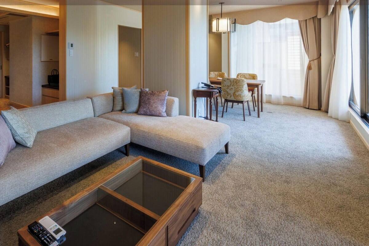 A modern hotel room with a large sectional sofa, a glass coffee table, a separate bed, a dining area with a table and chairs, and a large window with curtains.