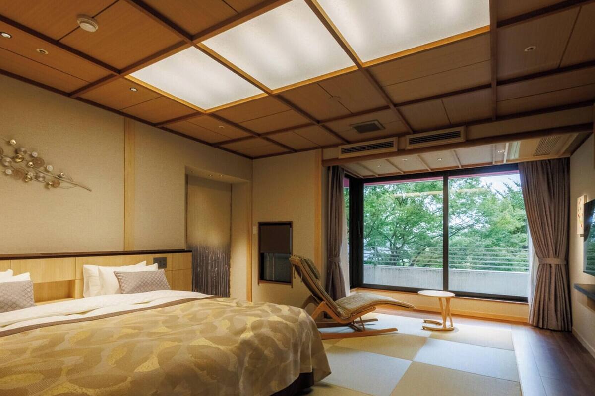 A hotel room with a large bed, a chair, a TV, and a window with a view of trees.