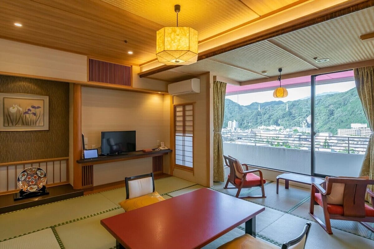 A traditional Japanese room with tatami flooring, a television, a dining table, and a balcony with a view of the city.