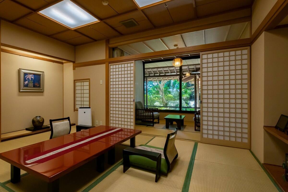 A traditional Japanese room with tatami flooring, a low table, chairs, a sliding door, and a view of a garden.