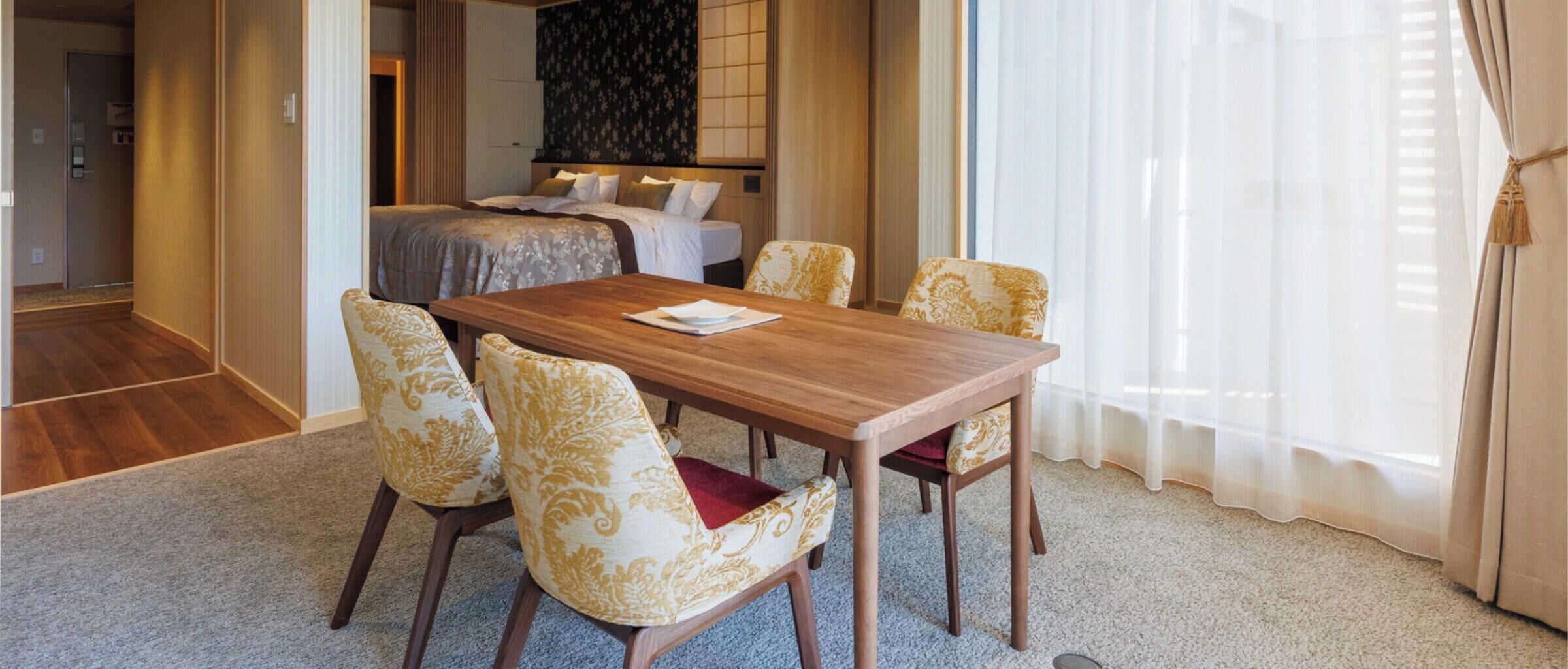 Habitación de hotel con mesa de comedor de madera, cuatro sillas estampadas, cama con cabecera floral y un ventanal con cortinas traslúcidas.