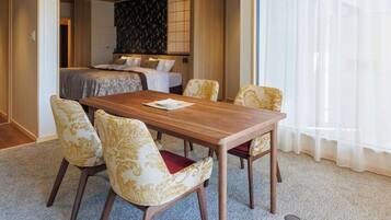 Habitación de hotel con mesa de comedor de madera, cuatro sillas estampadas, cama con cabecera floral y un ventanal con cortinas traslúcidas.