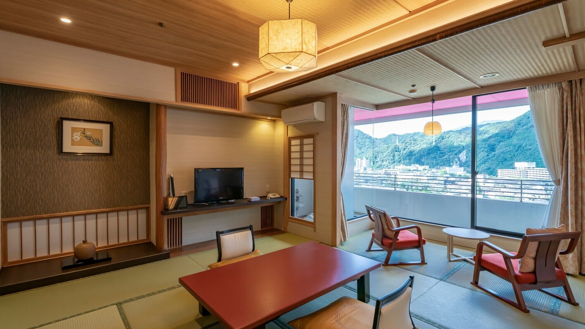 A hotel room with a TV, a dining table, chairs, a large window with a view, and traditional decor.
