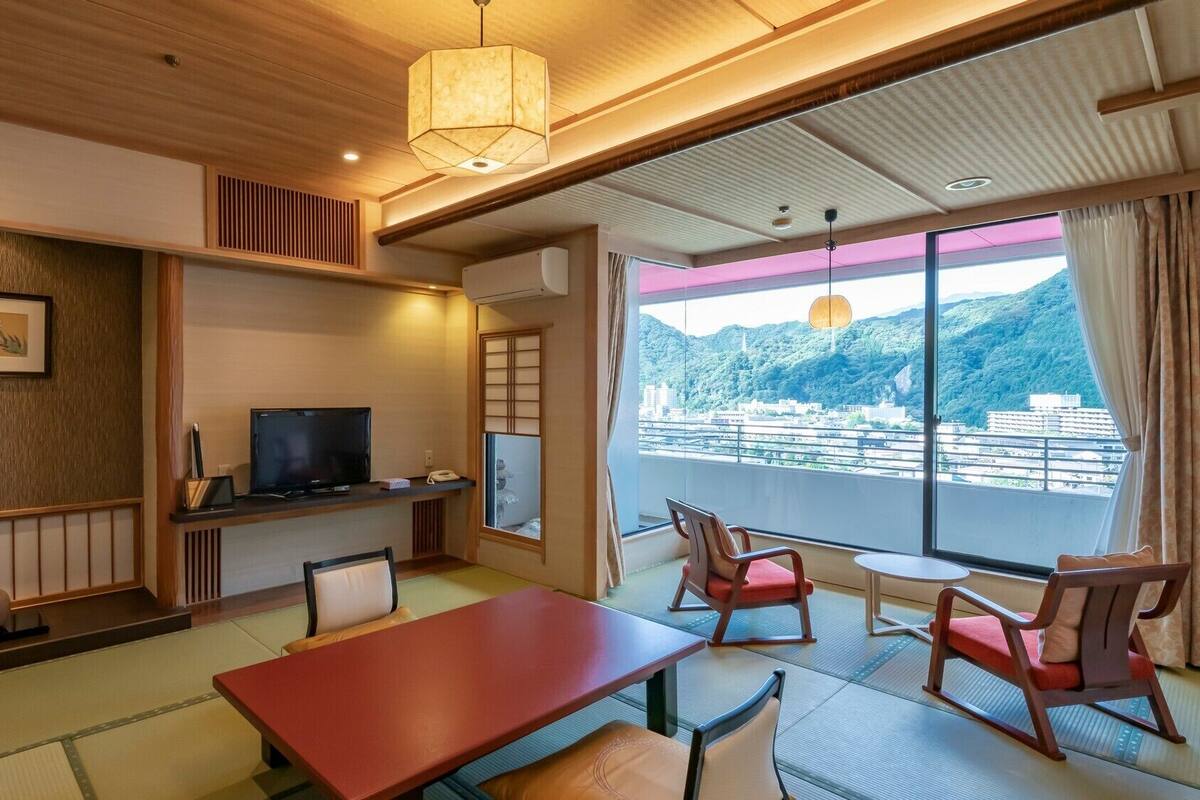 A hotel room with a TV, a dining table, chairs, a large window with a view, and traditional decor.