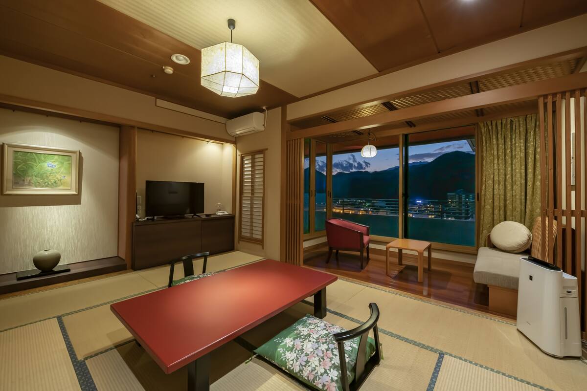 A traditional Japanese-style room with tatami flooring, a low table, a TV, and a view of a cityscape through large windows.