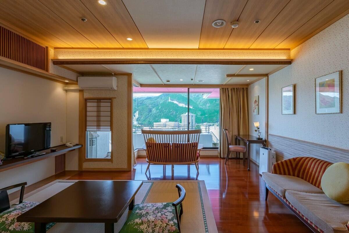 A hotel room with a balcony, a TV, a sofa, a dining table, and a chair.