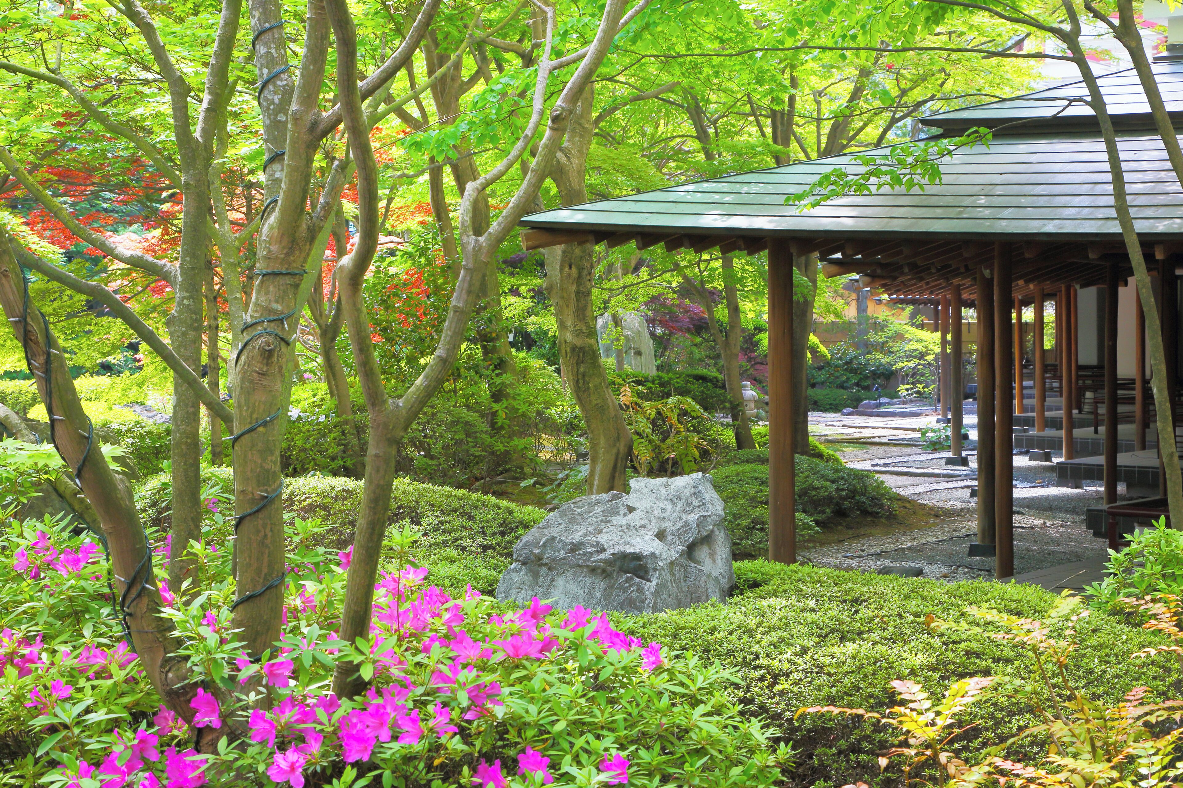 A traditional Japanese garden with a wooden pavilion, lush greenery, and a large rock centerpiece.