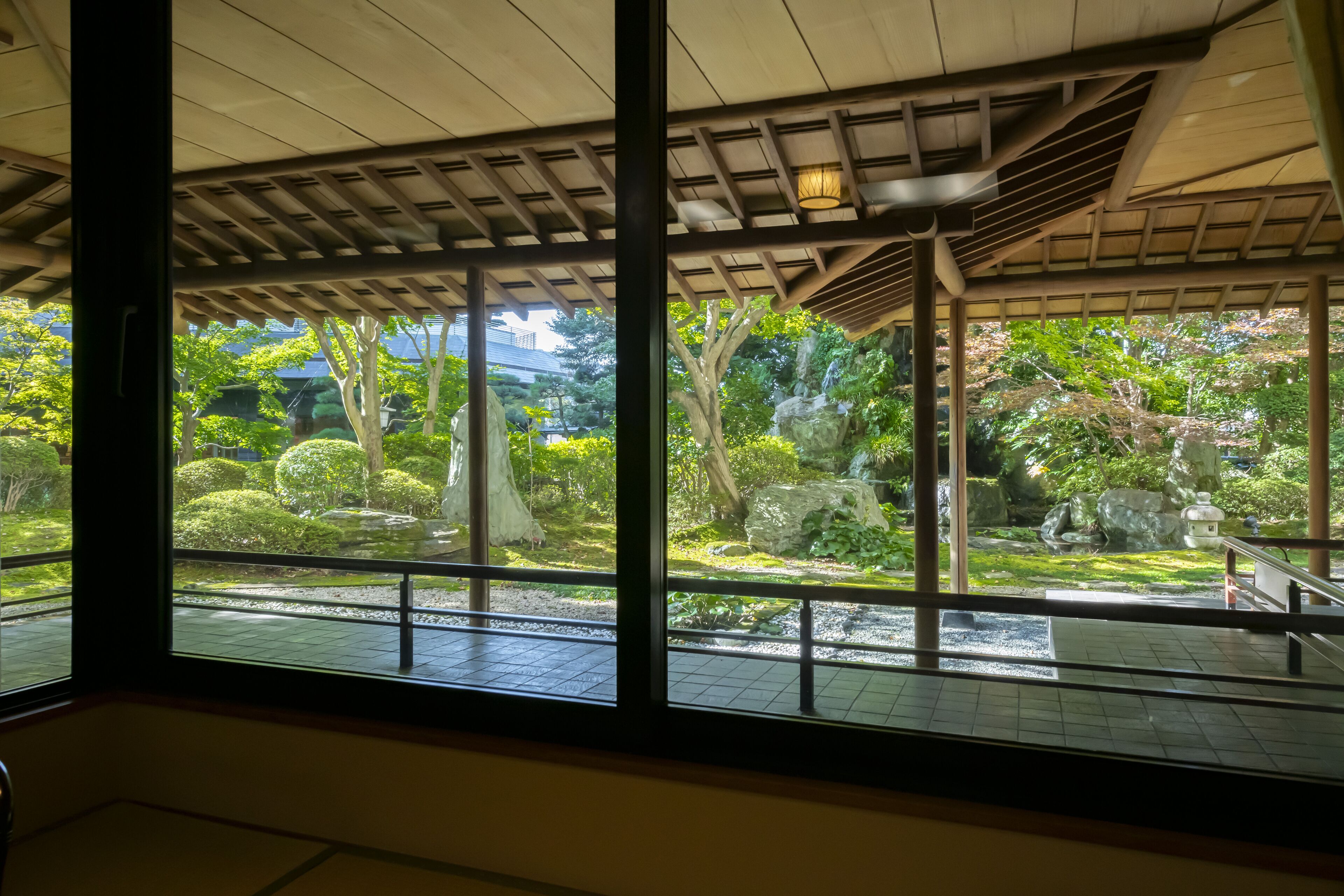 Un interior japonés tradicional con grandes ventanales que ofrecen vistas a un jardín con rocas, árboles y una pequeña cascada.