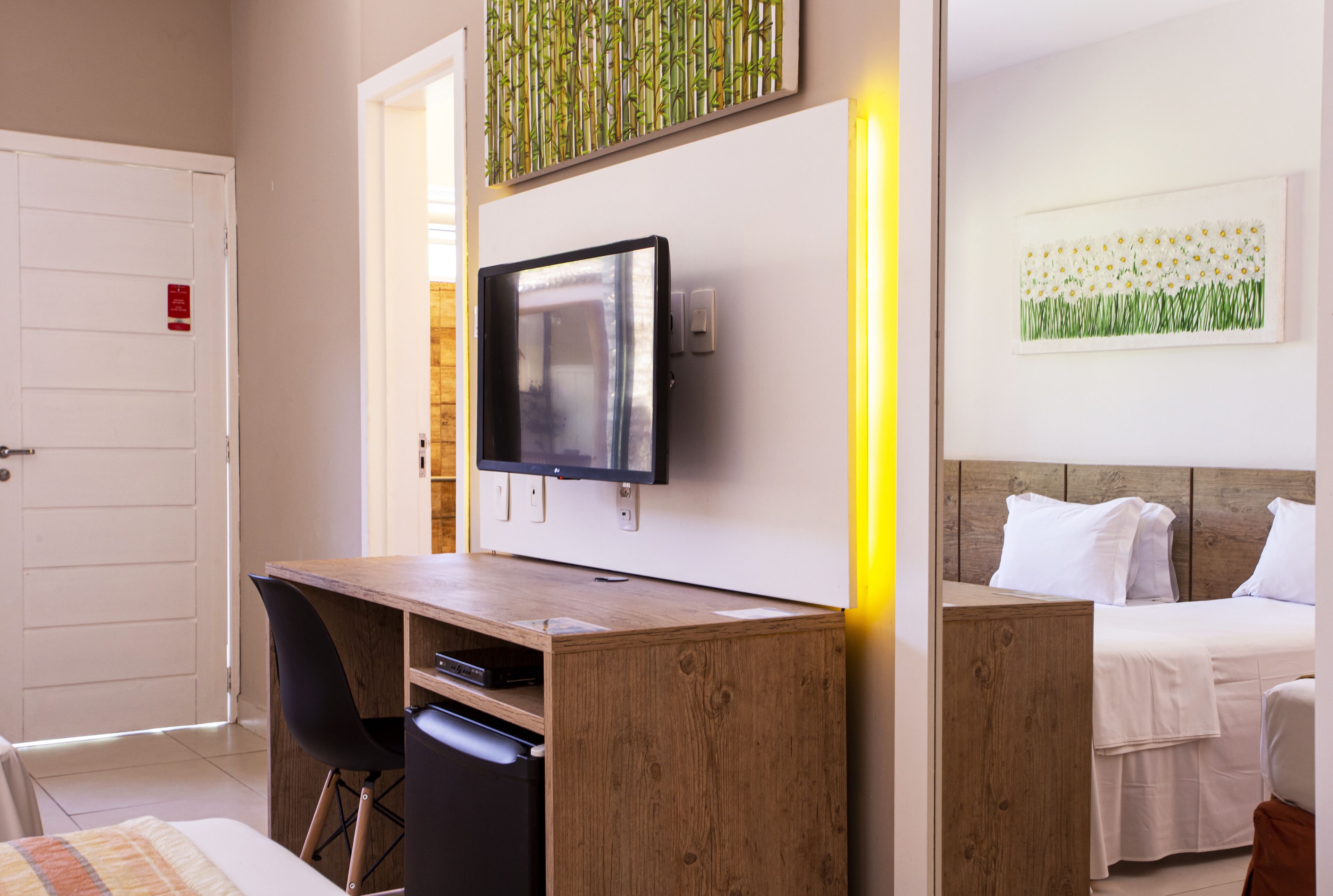 A hotel room with a wooden desk, a television, a bed with white bedding, and a wall-mounted artwork of flowers.