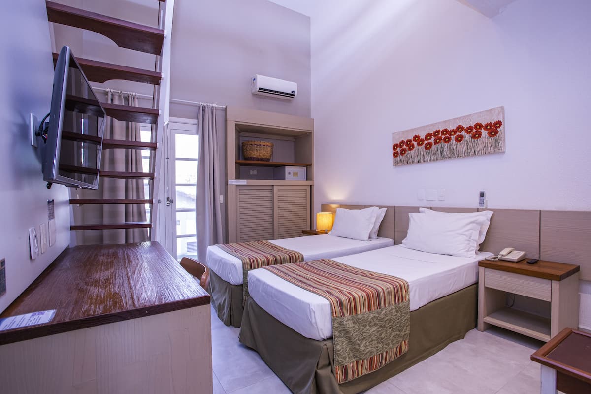 A hotel room with two beds, a TV mounted on the wall, an air conditioning unit, a bedside lamp, and a decorative wall piece.