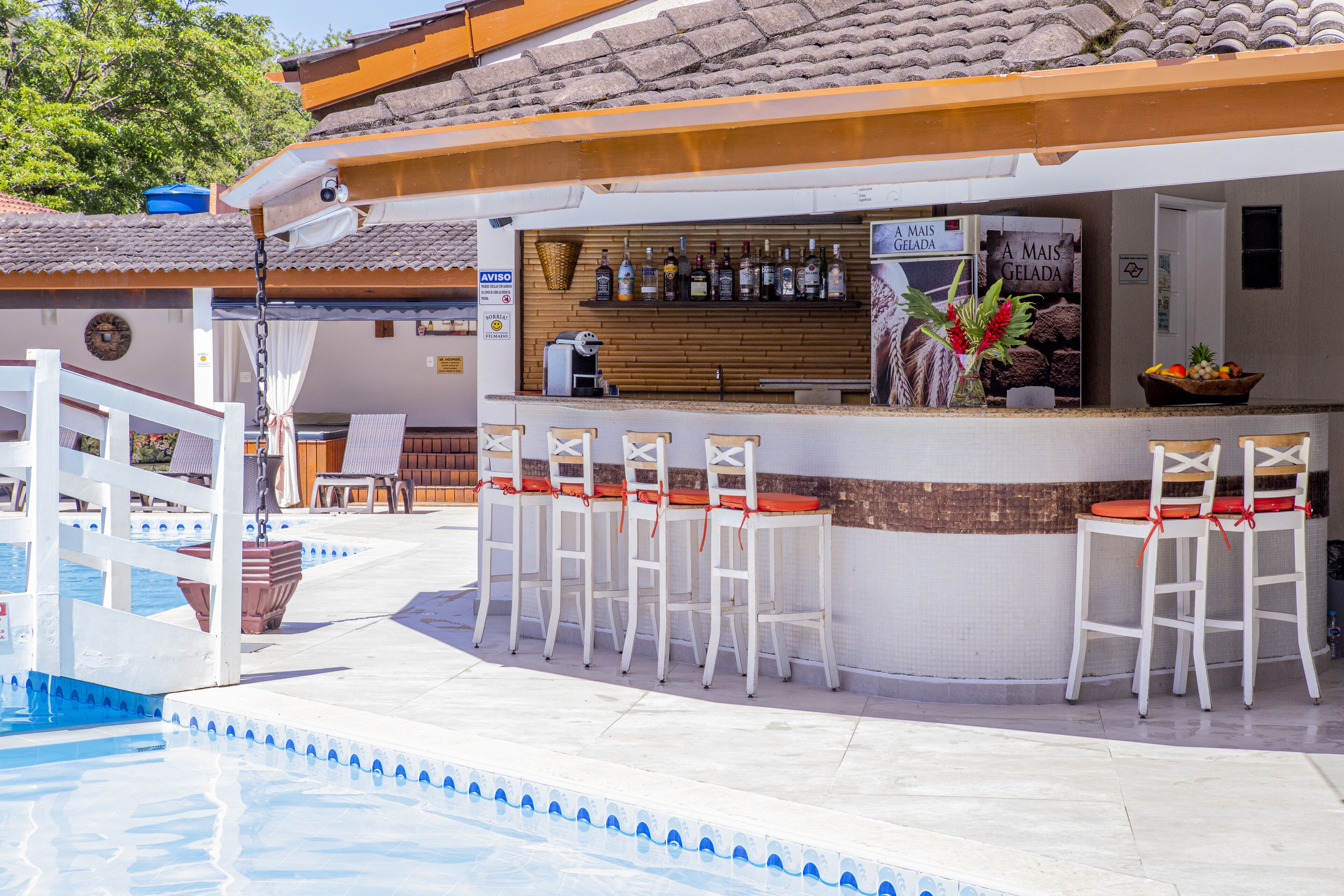 A bar area with a poolside seating arrangement, a bar counter with a variety of bottles, and a sign that reads "A MAIS GELADA".