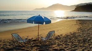 A beach with two beach chairs and a blue umbrella labeled "CIRIBAI" under a sunset. - Ciribaí Praia Hotel (São Sebastião)