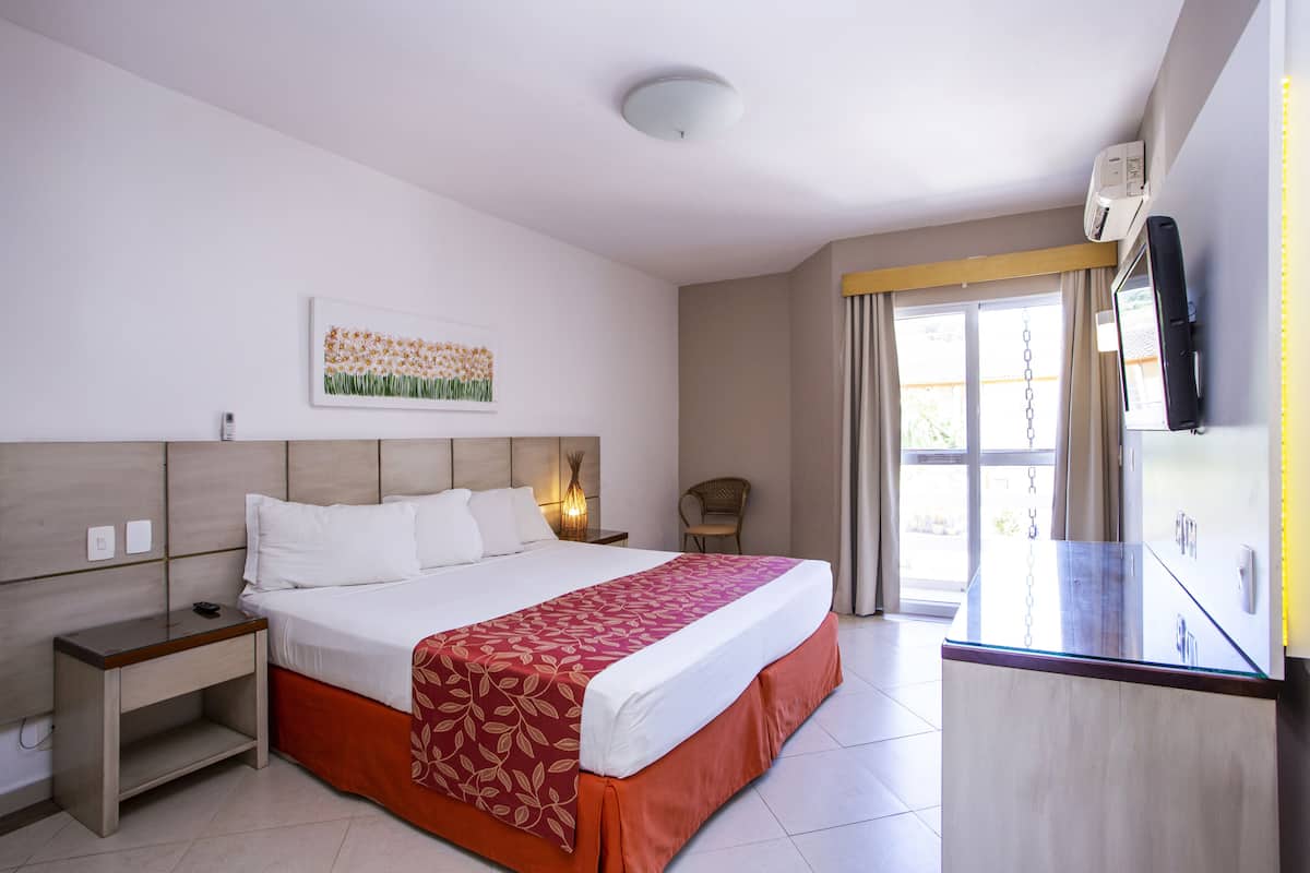 A hotel room with a bed, bedside table, nightstand, wall art, and a window with curtains.