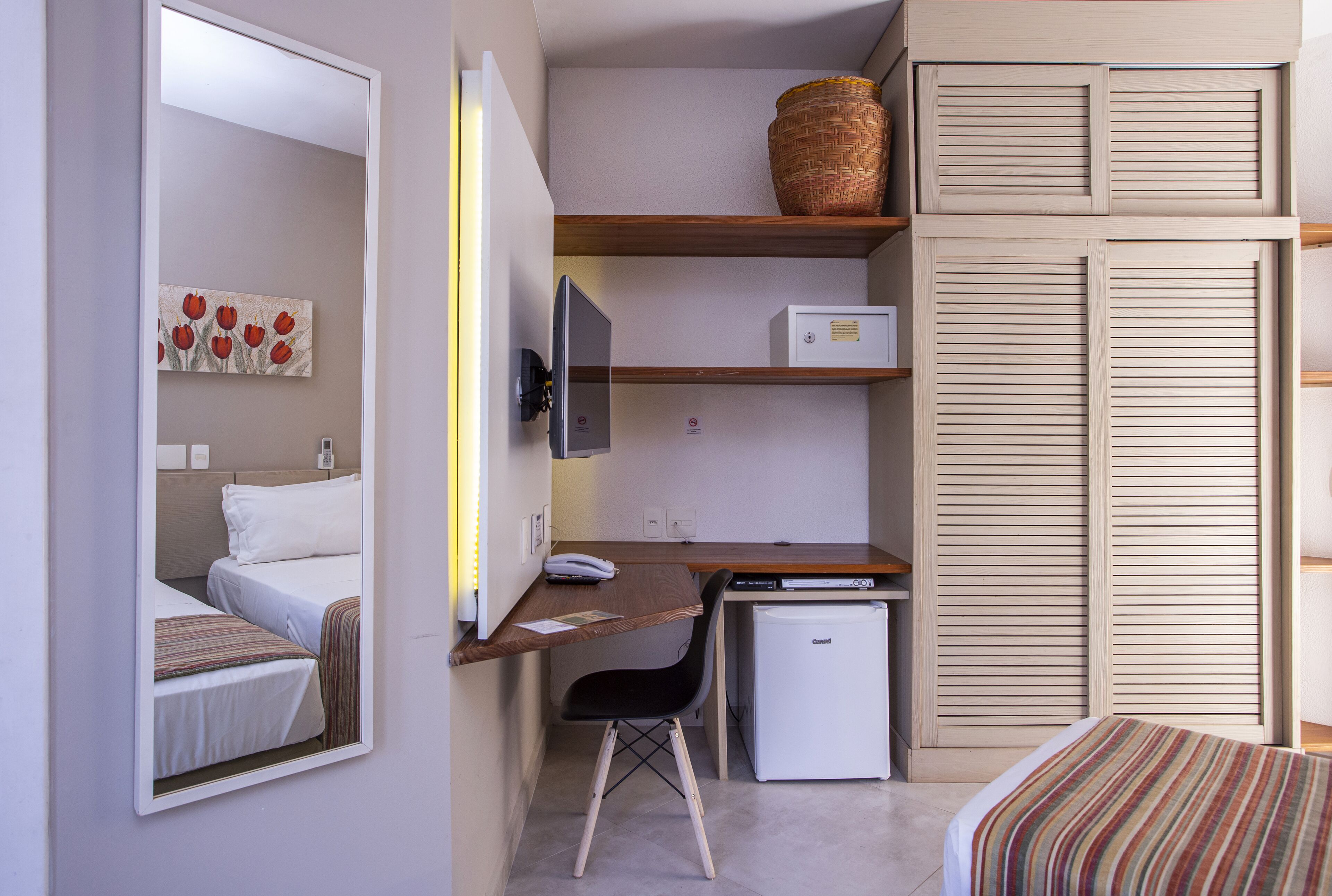 Een knus hotelkamer met een bed, een bureau met computer, een kleine koelkast, een stoel en een spiegel.