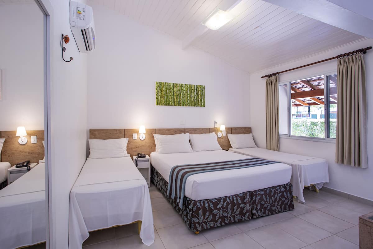 A hotel room with two beds, a wooden headboard, a wall-mounted air conditioner, and a window with curtains.