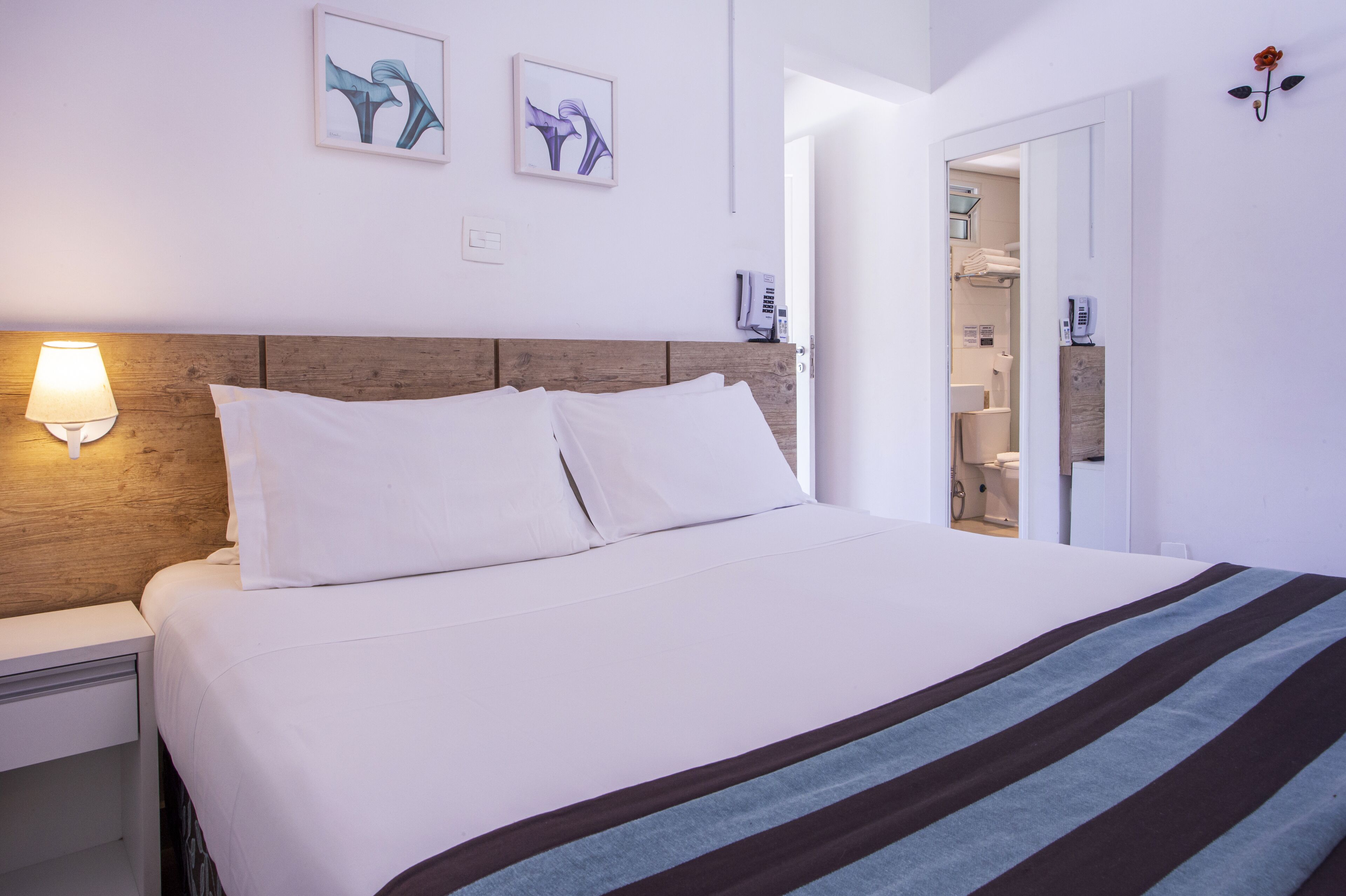 Quarto de hotel com cama, mesa de cabeceira, luminária na parede e duas obras de arte emolduradas.