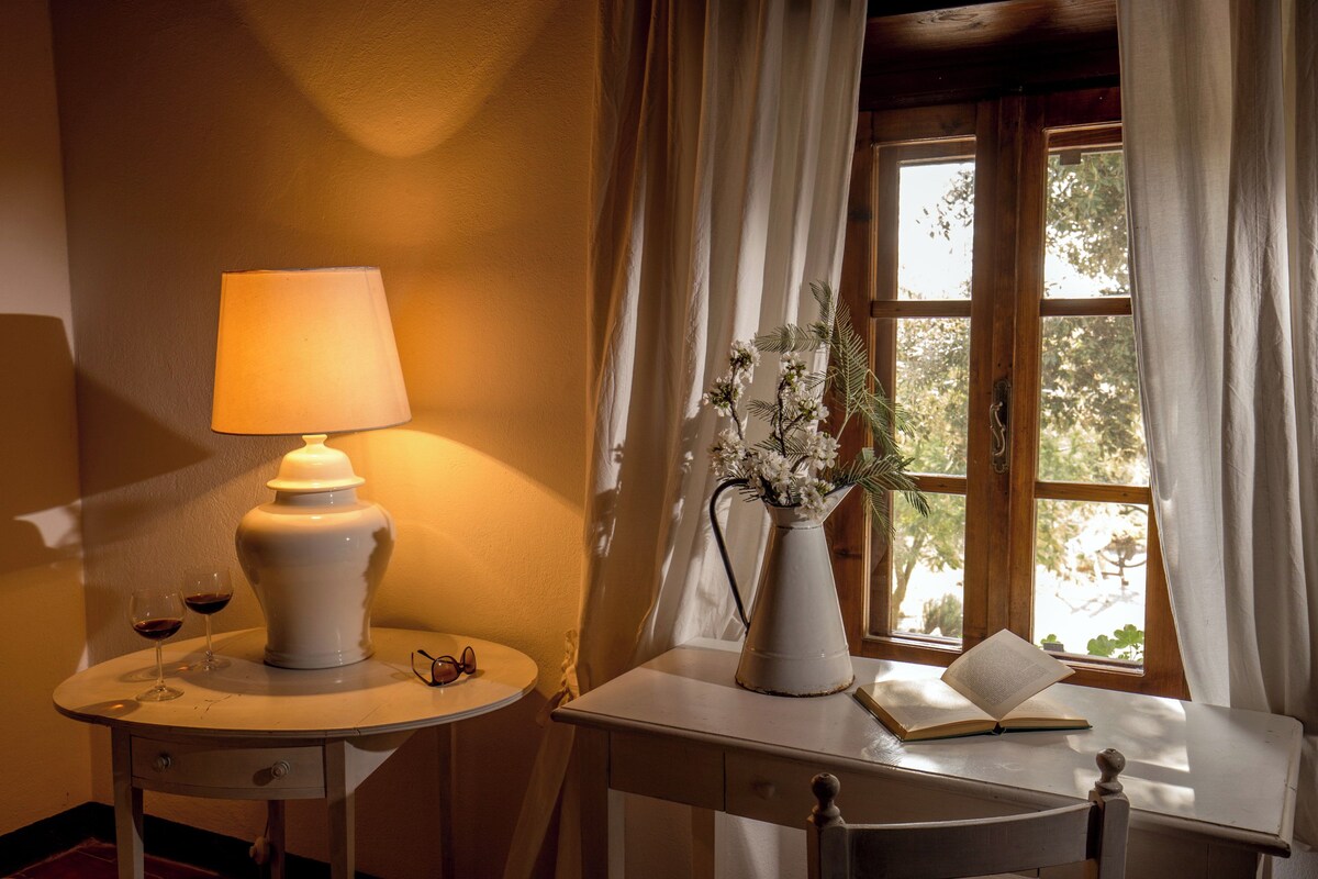 A room with a table, a lamp, two wine glasses, a vase with flowers, and an open book.