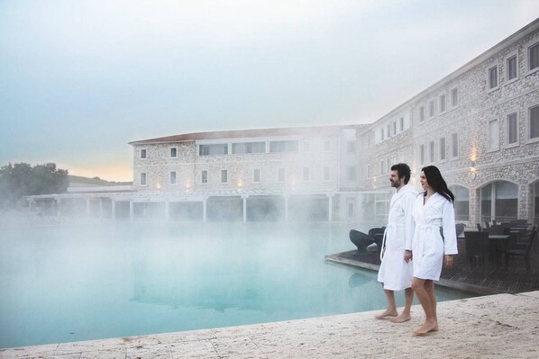 Una pareja parada junto a una piscina con vapor saliendo del agua, frente a un edificio de piedra con varias ventanas.