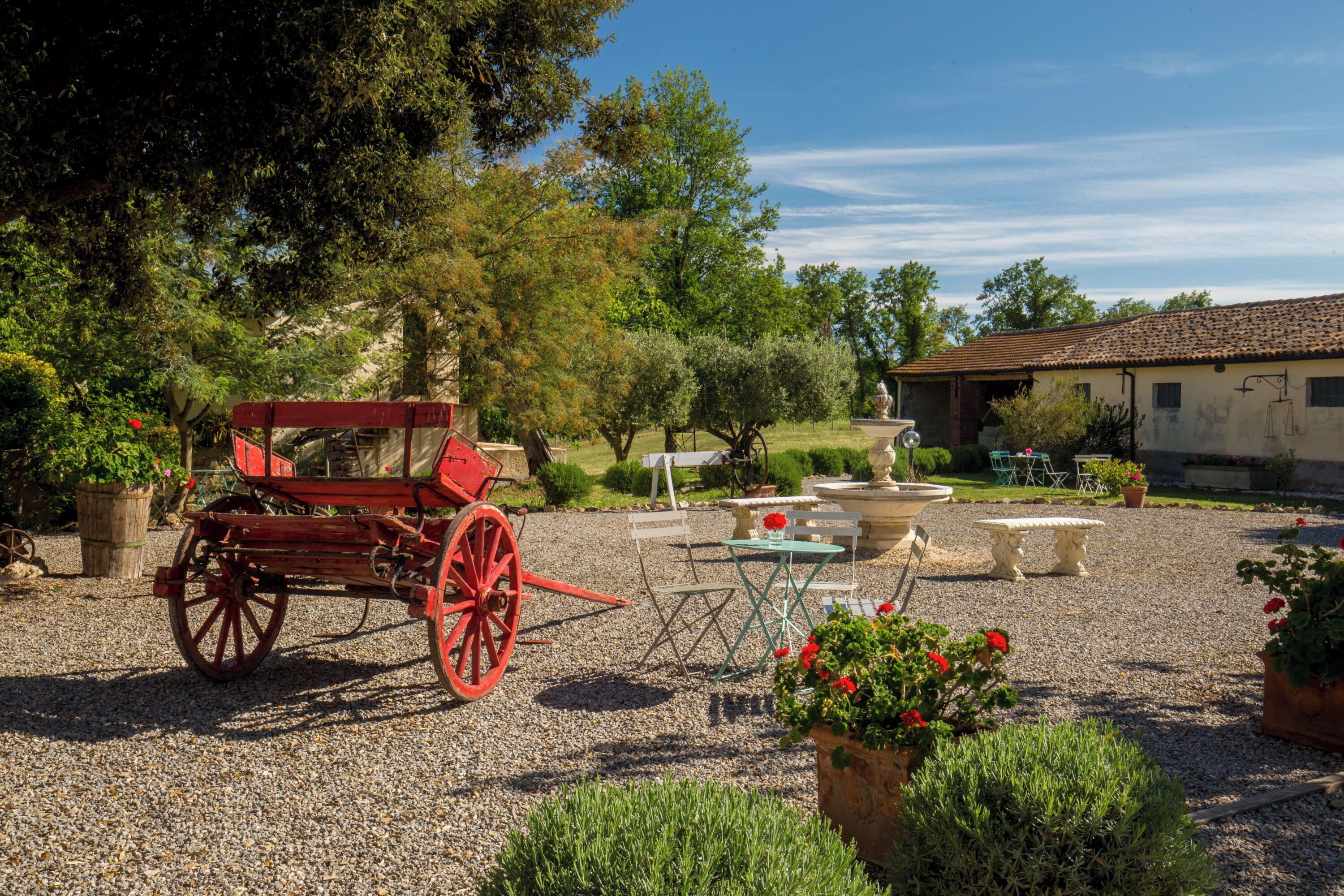 A rustic outdoor setting with a red carriage, gravel ground, potted plants, and outdoor furniture.