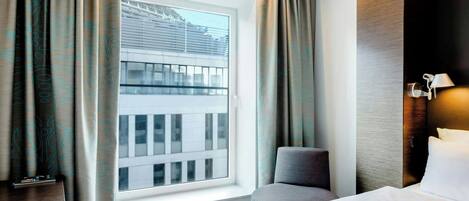 A hotel room with a bed, a desk, a chair, and a window with a view of a building.