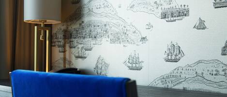 A room with a patterned wallpaper depicting ships and coastal scenes, a blue chair, and a lamp with a white shade.