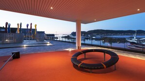Un espacio exterior con techo de madera, una zona de estar circular y vistas a un puerto con barcos.