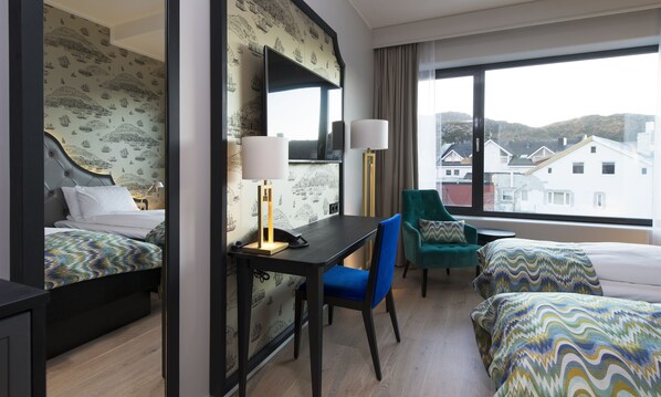 A hotel room with two beds, a desk with a lamp, a chair, and a large window with a view of buildings. - Thon Hotel Fosnavag (Heroy)