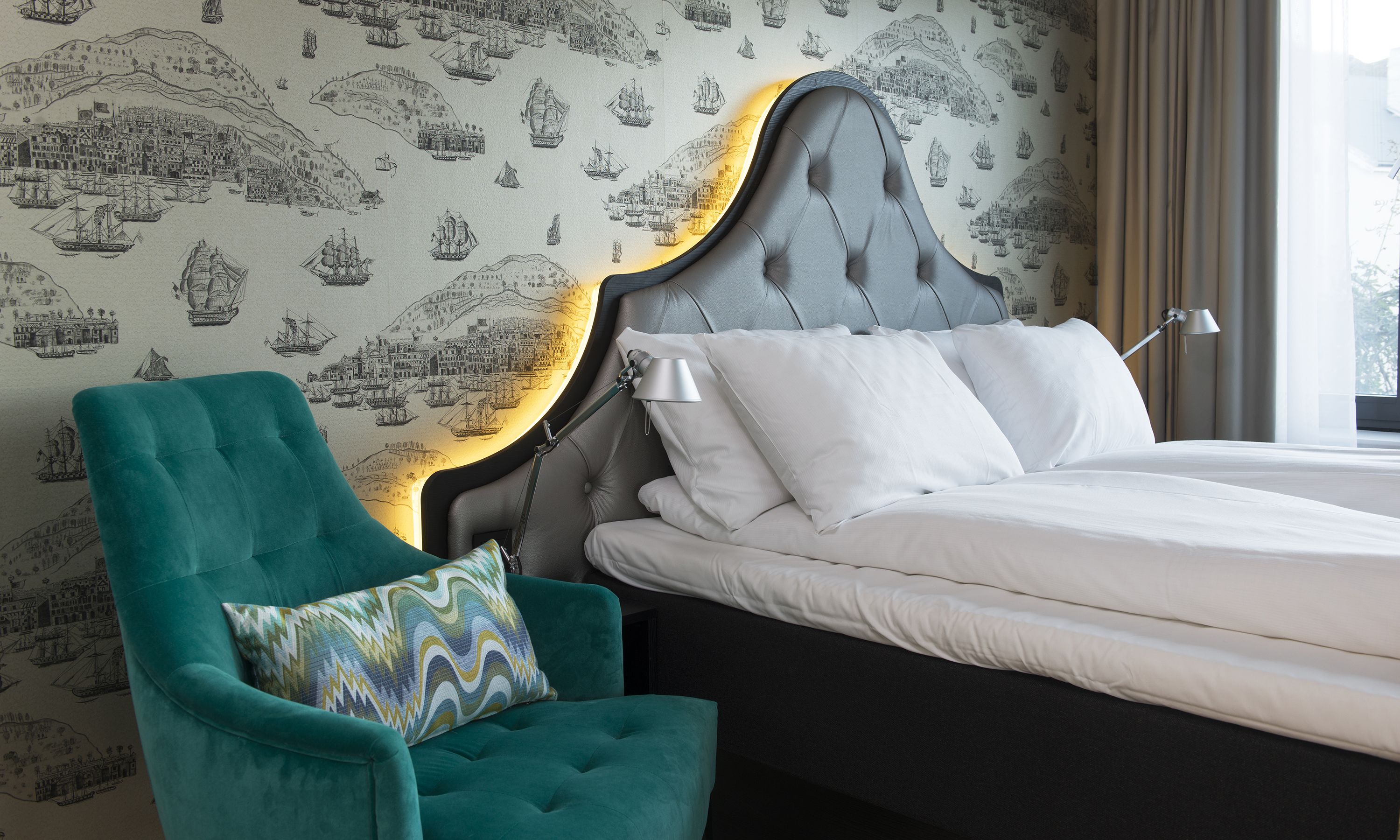A neatly made bed with white linens, a teal armchair with a patterned cushion, and a wall with maritime-themed wallpaper.