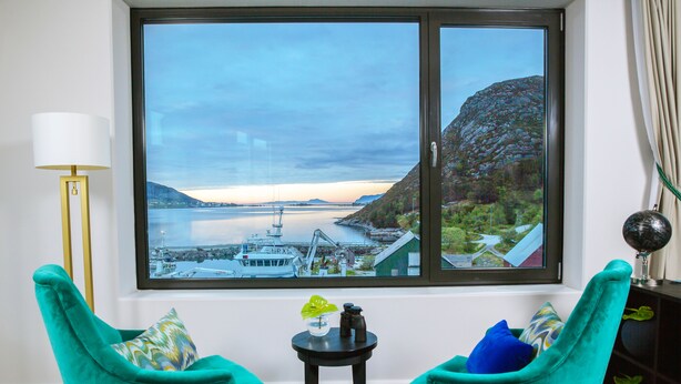 Habitación con un amplio ventanal que ofrece vistas a una masa de agua y un paisaje montañoso. Hay dos sillas color turquesa y una mesita con adornos.